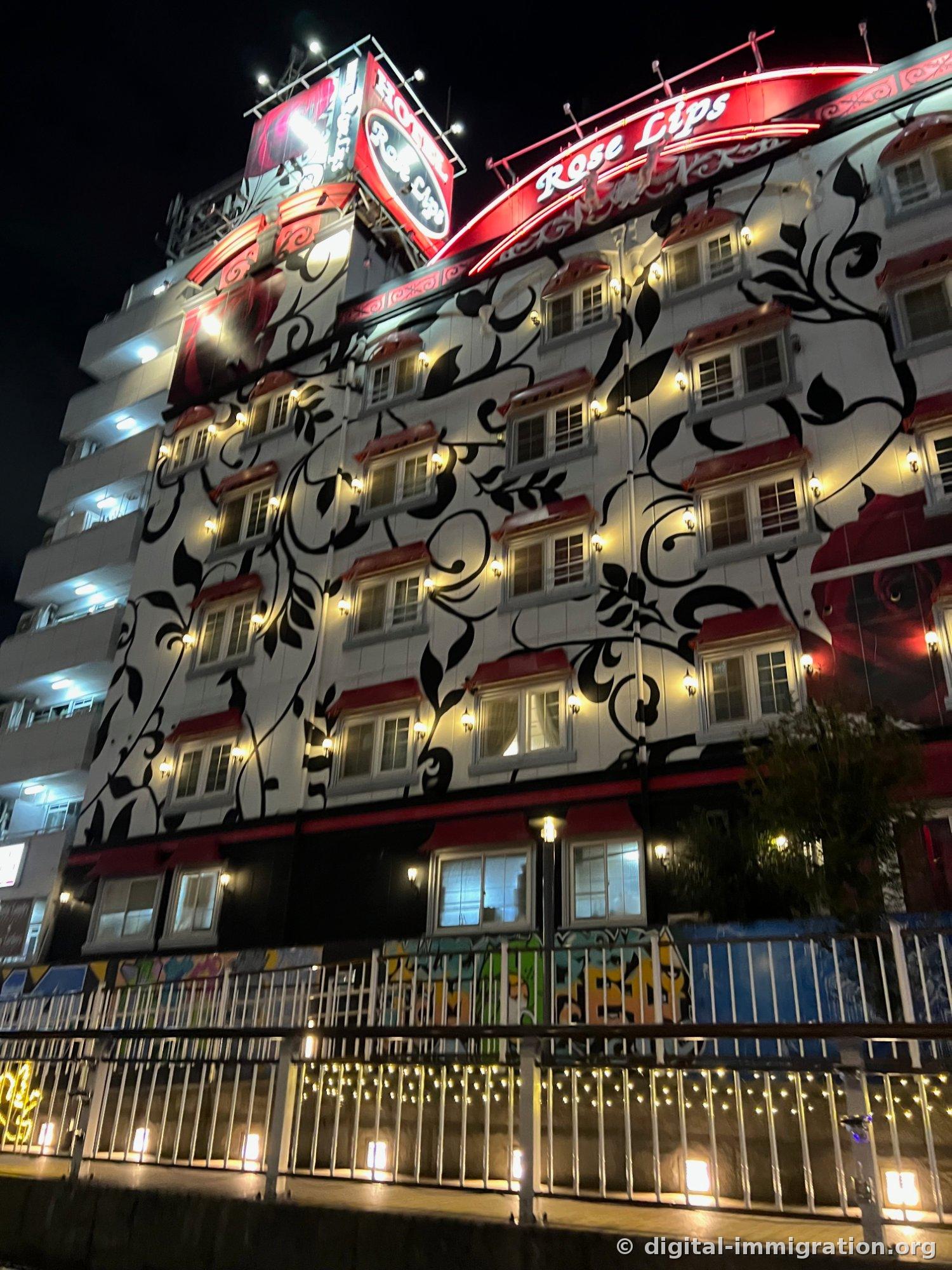 Façade extérieure d'un bâtiment blanc couverte de motifs de roses grimpantes et enseigne néon rouge 'Rose Lips' accrochée à un hôtel la nuit