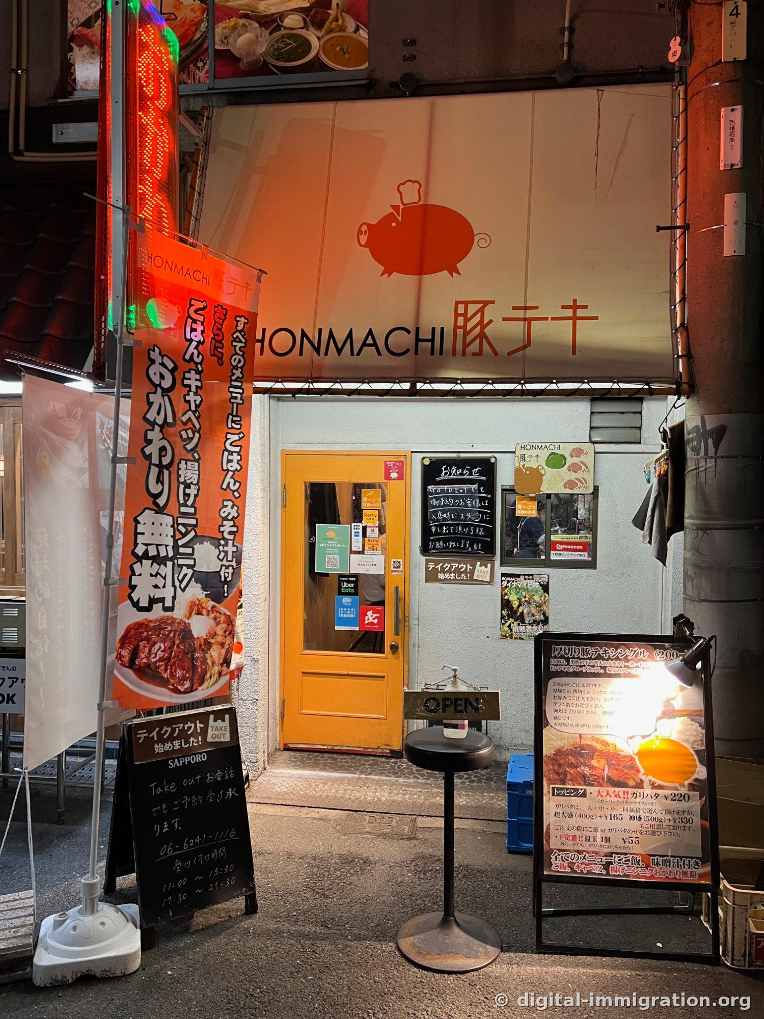 The entrance of HONMACHI 豚テキ photographed at night. A sun awning with a pink pig logo, a yellow sliding door, an orange nobori flag on the left with text about free refills and a chalkboard with takeout information, and a standing sign on the right with the tonkateki menu.