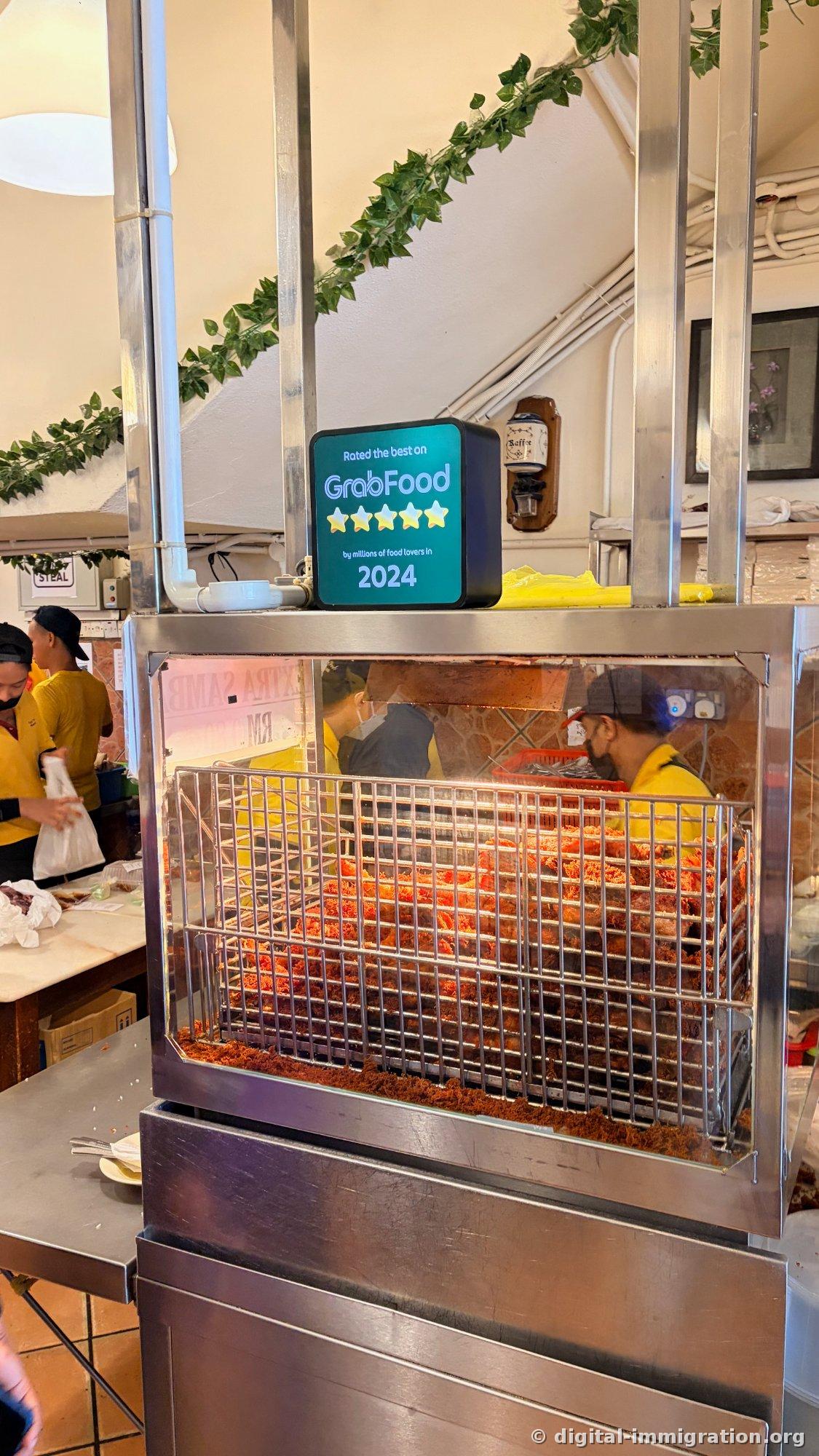 Inside a stainless steel warming display, fried chicken in reddish sauce is stacked in layers, fried crumbs coat the bottom, and a 'Rated the best on GrabFood 2024' light box sits on top, with staff in yellow uniforms on the left continuing to pack takeout orders