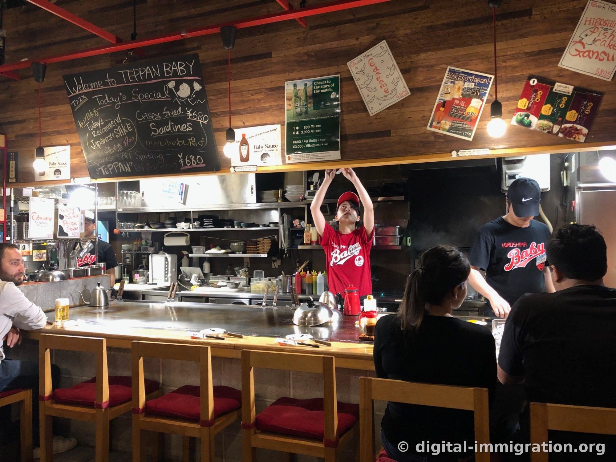 붉은 철골 천장 아래 긴 철판 카운터와 Welcome to TEPPAN BABY 칠판, 붉은 히로시마 베이비 티셔츠를 입은 직원