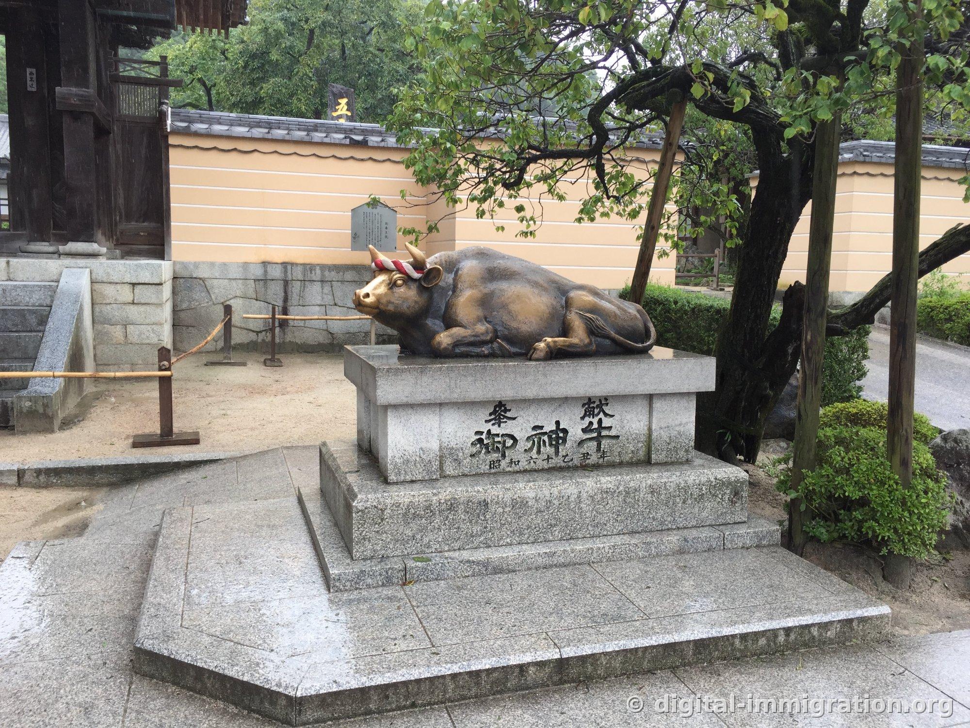 Bronze cow statue lying on a granite pedestal with the inscription '奉献 御神牛' and fence in the background
