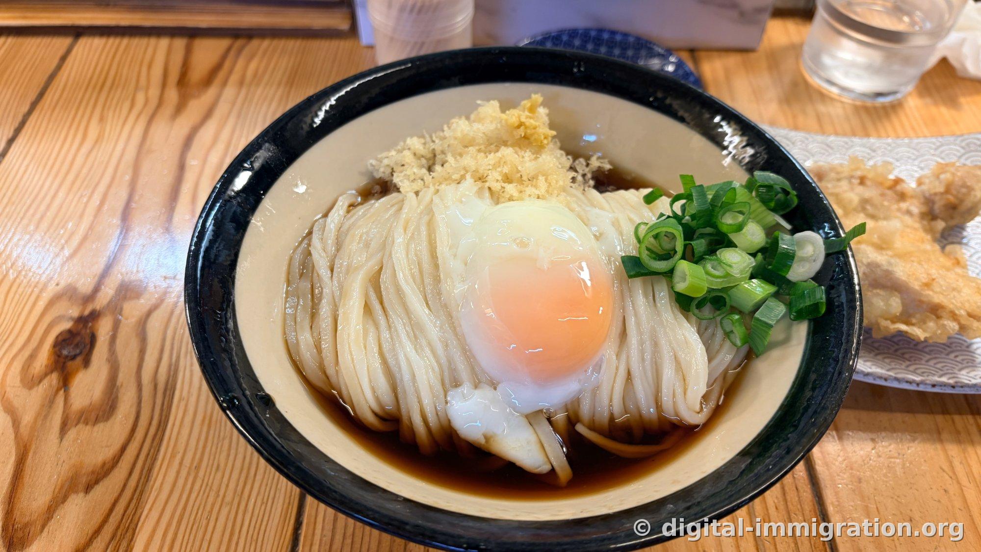 黒い縁取りの丼に盛られたぶっかけうどん。濃いめの醤油だしにとろろ卵の黄身、天かす、すりおろし生姜、小口切りのネギが載っており、脇に天ぷら皿が置かれている