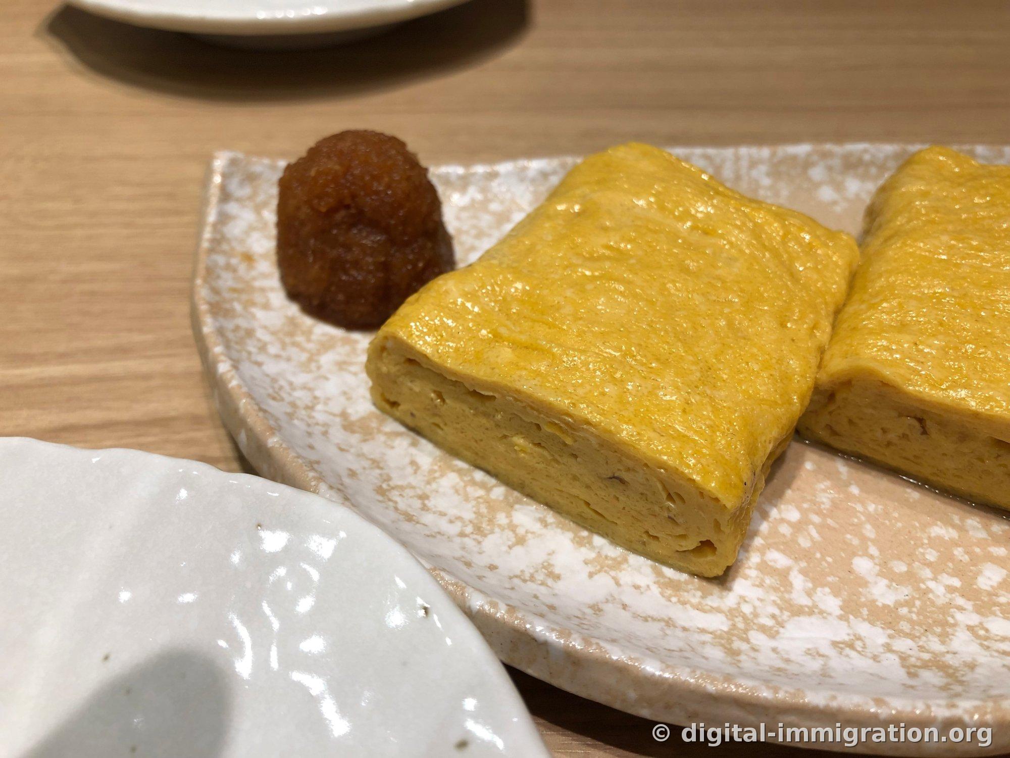 A close-up of thick yellow tamagoyaki sections on a white glazed plate and grated brown daikon bundled beside