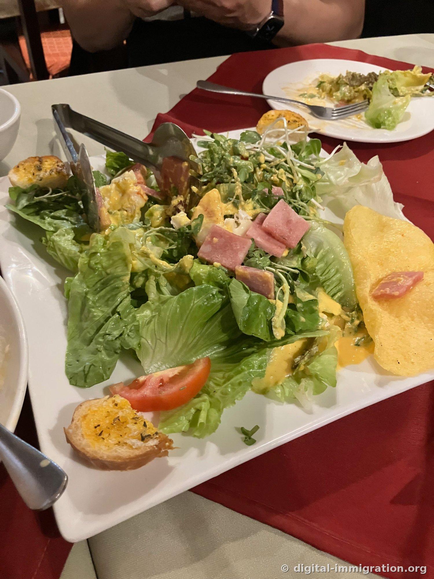 Green salad on a rectangular white plate with radish sprouts, diced ham, and yellow mustard dressing, accompanied by cheese-topped baguette slices and a large shrimp chip