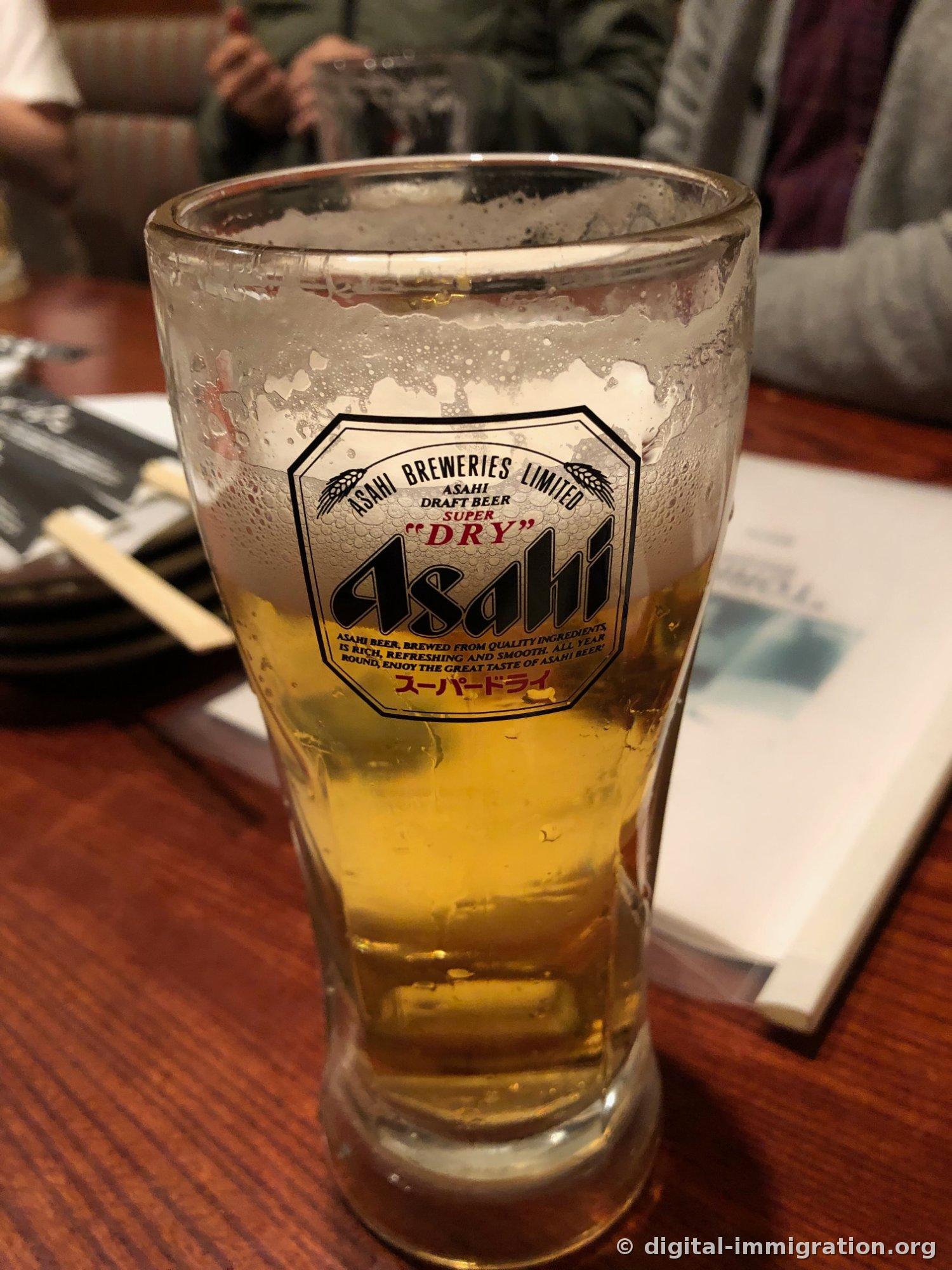 朝日超爽标志印在玻璃杯上，啤酒泡沫厚厚地堆积在杯口，木制桌面上放着木筷和菜单