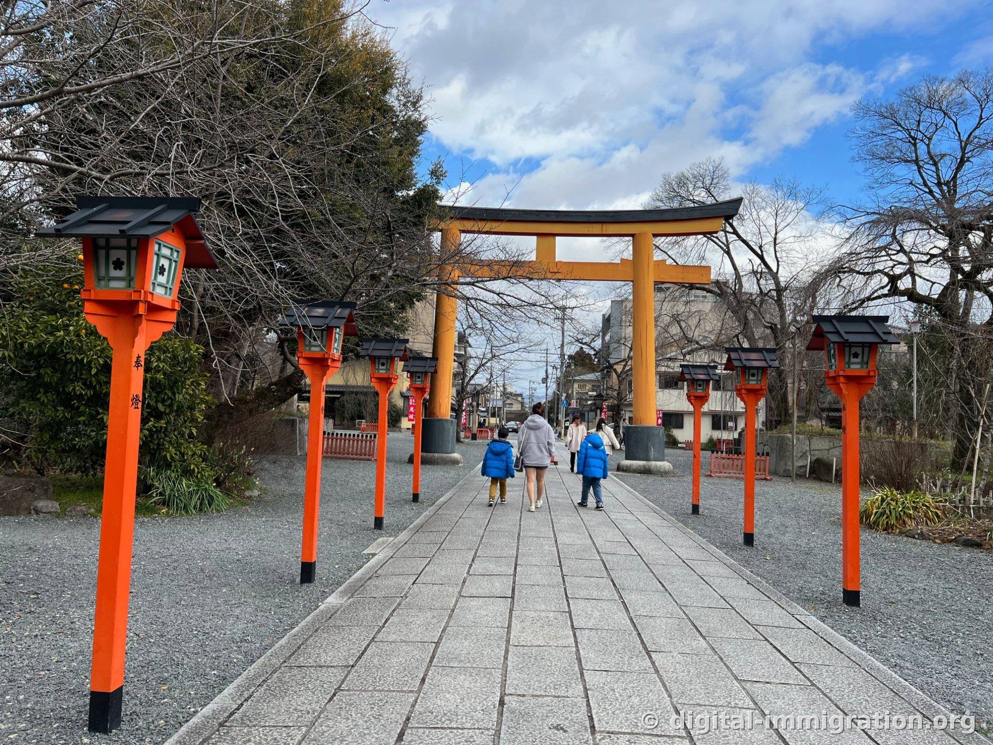 The southern approach path to Hirano Shrine leading toward a large orange torii gate, with red lantern pillars inscribed with '奉燈' lining both sides of the stone path