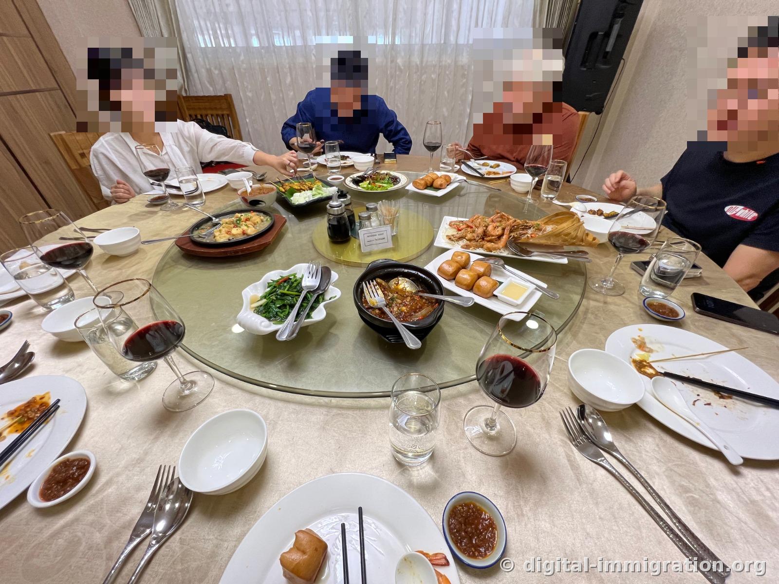 A glass lazy Susan laden with iron plate tofu, satay skewers, large prawns, stir-fried water spinach, fried mantou with condensed milk, and a clay pot of spicy sauce, with diners enjoying red wine