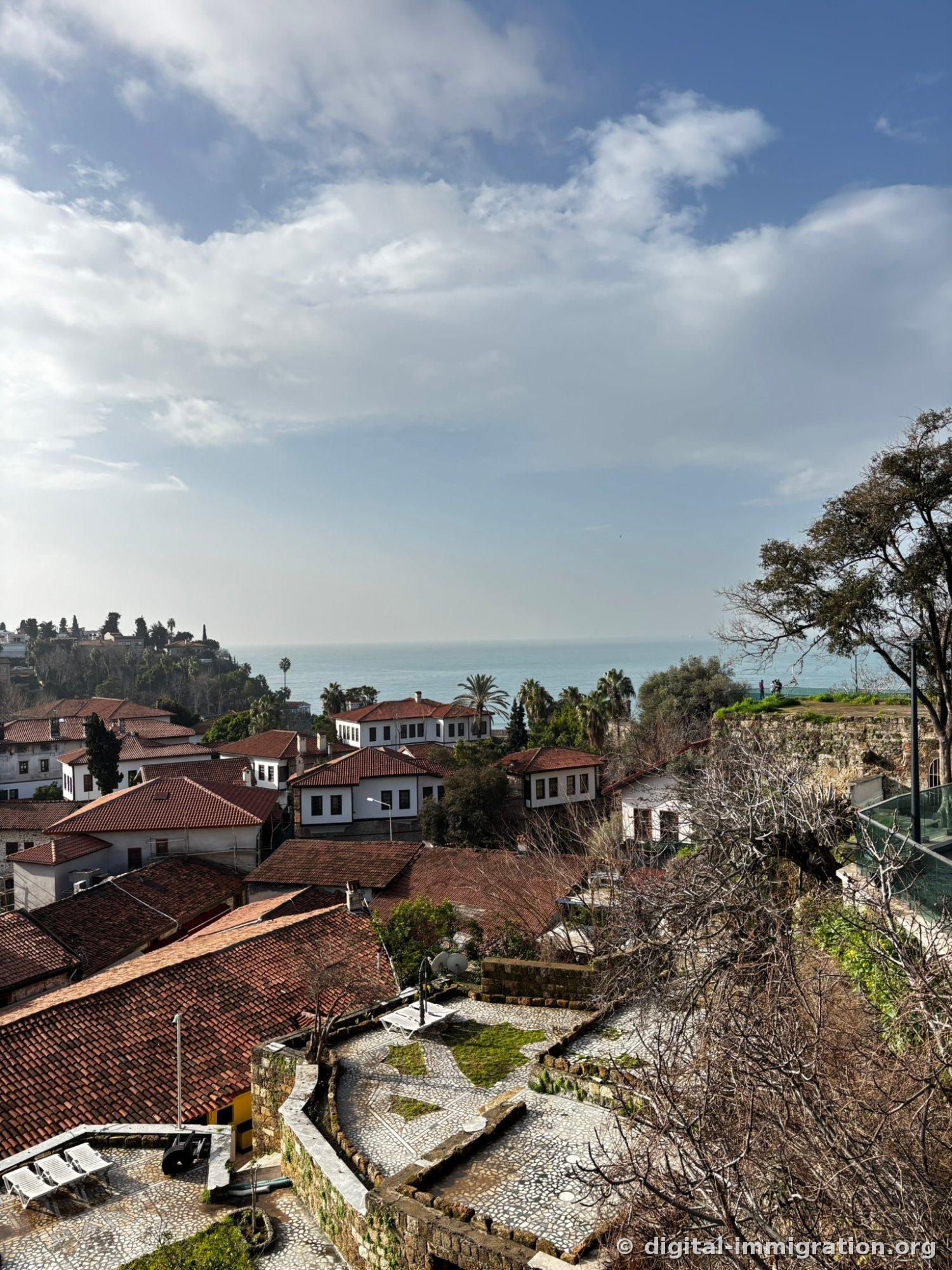 Looking down from the cliff at the red tile roofs of the old city of Kaleiçi and white Ottoman houses, with the Mediterranean horizon and palm trees stretching beyond.