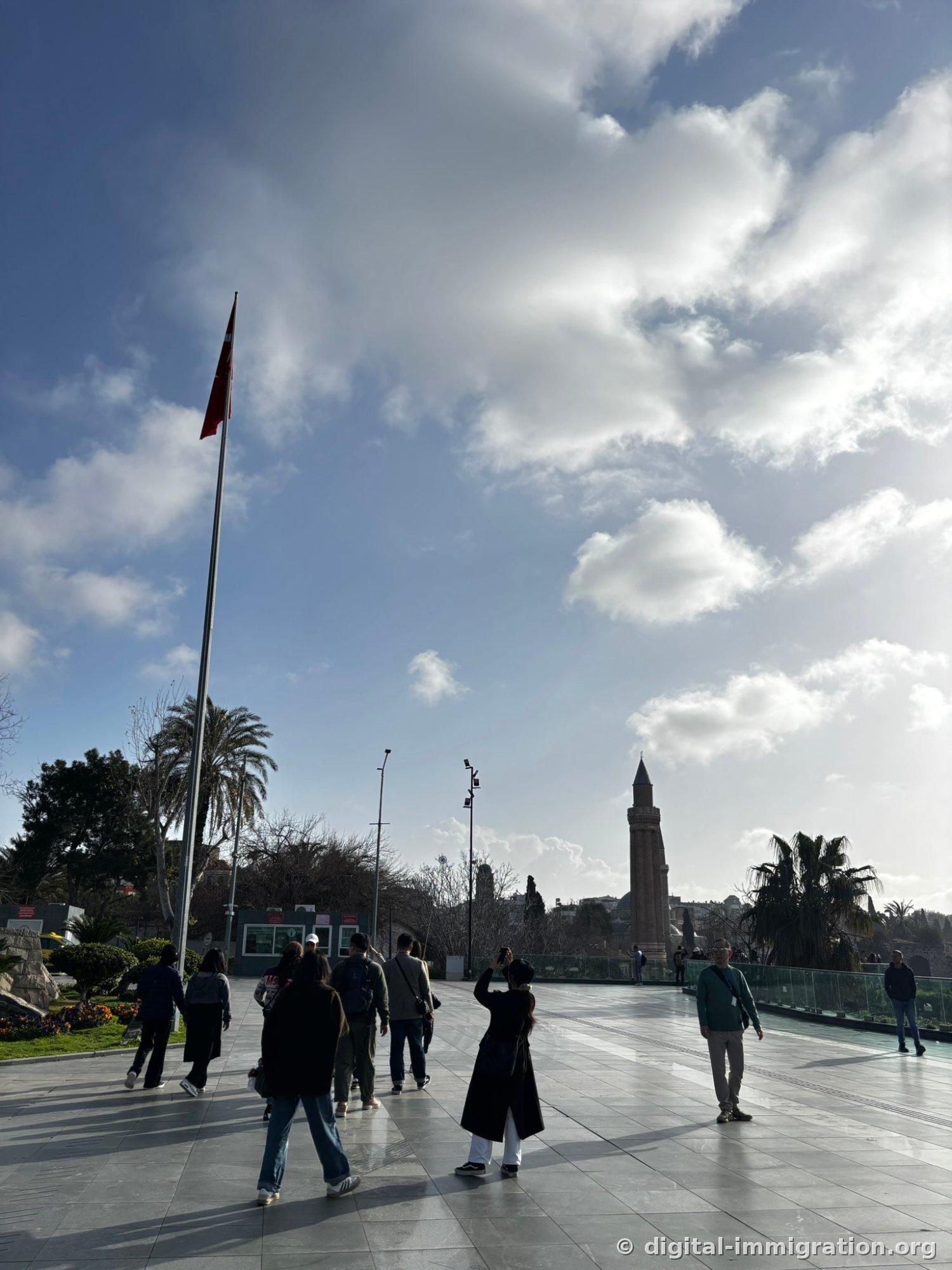 People walking across a broad marble square, a tall flag pole and palm tree on the left, the red brick spire of the Yivli Minaret standing far right against a cloudy sky.