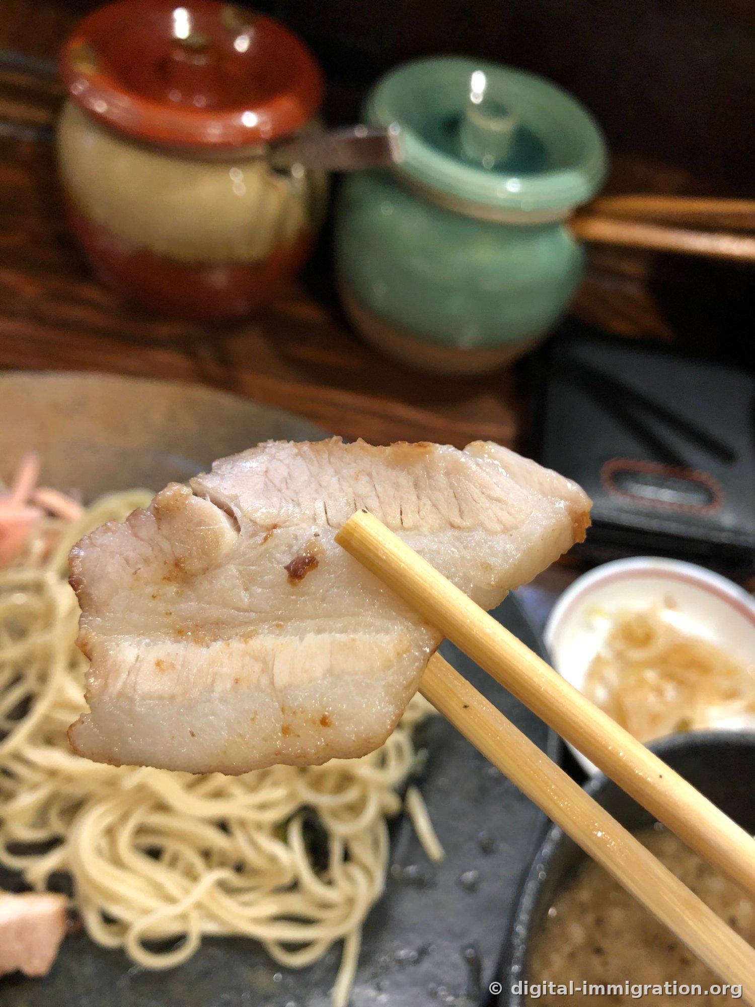 Une tranche épaisse de char siu de poitrine saisie avec des baguettes. La couche de graisse est cuisonnée à la perfection, semi-transparente, avec l'assiette de nouilles et le bol de bouillon en dessous
