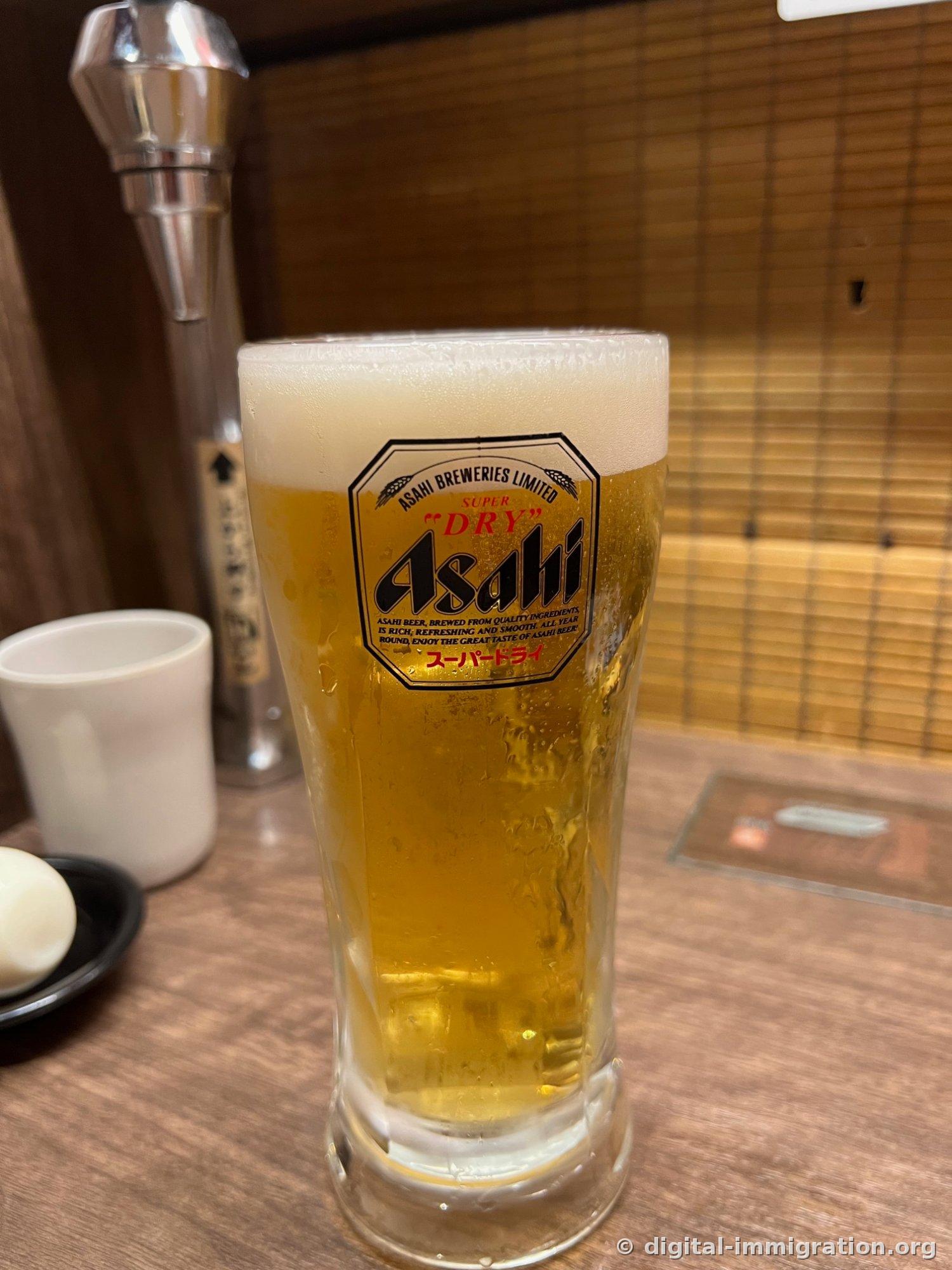 A frosted glass of Asahi Super Dry draft beer sitting on the wooden counter of a booth seat, with self-serve water dispenser and bamboo footrest visible in the back