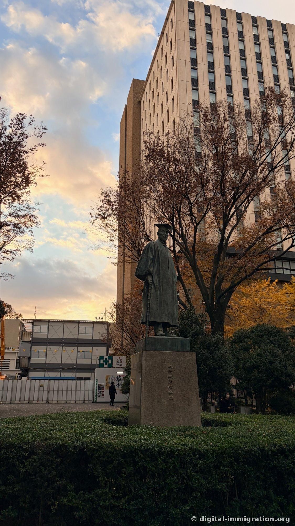 日落天空下穿着学位服站立的青铜雕像和后面的现代建筑，施工挡板