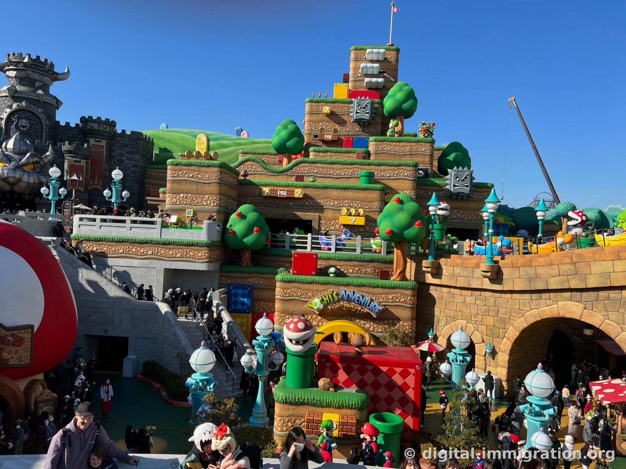 Overview of Super Nintendo World with question mark blocks, POW blocks, apple trees, and stone face characters arranged on tiered hills, with the gray Bowser's Castle on the left