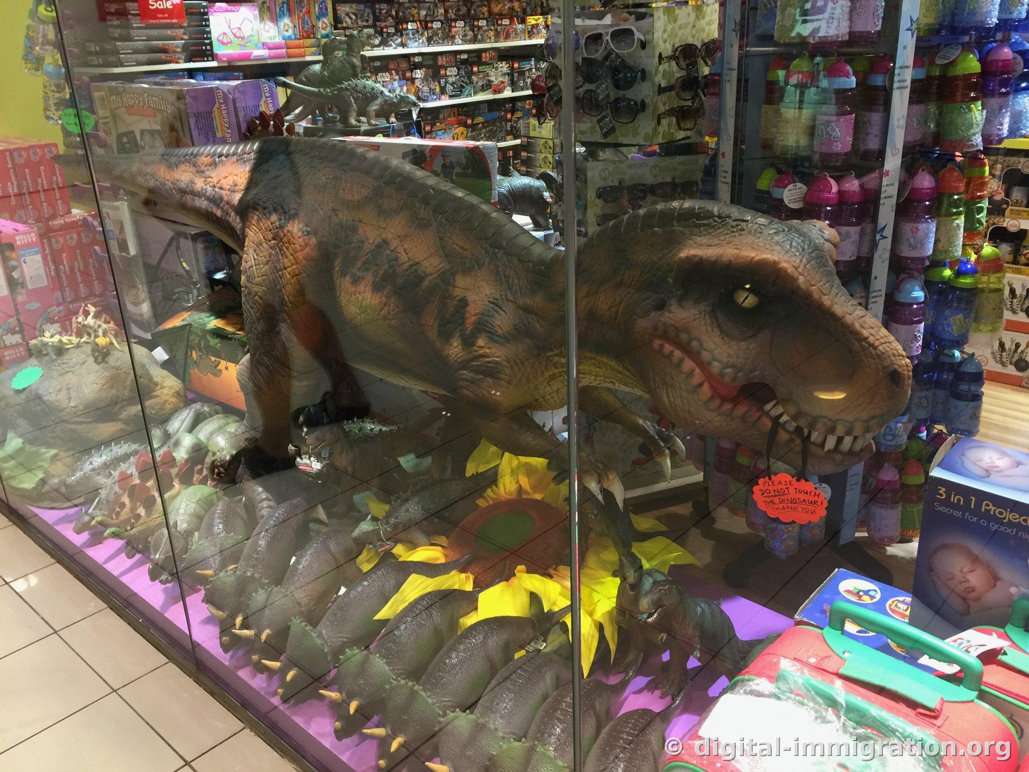 Un grand modèle de Tyranosaure Rex dans une vitrine en verre. Une pancarte écrite à la main « PLEASE DO NOT TOUCH THE DINOSAUR » est accrochée autour de son cou, avec des Lego et des bouteilles d'eau pour enfants affichées derrière