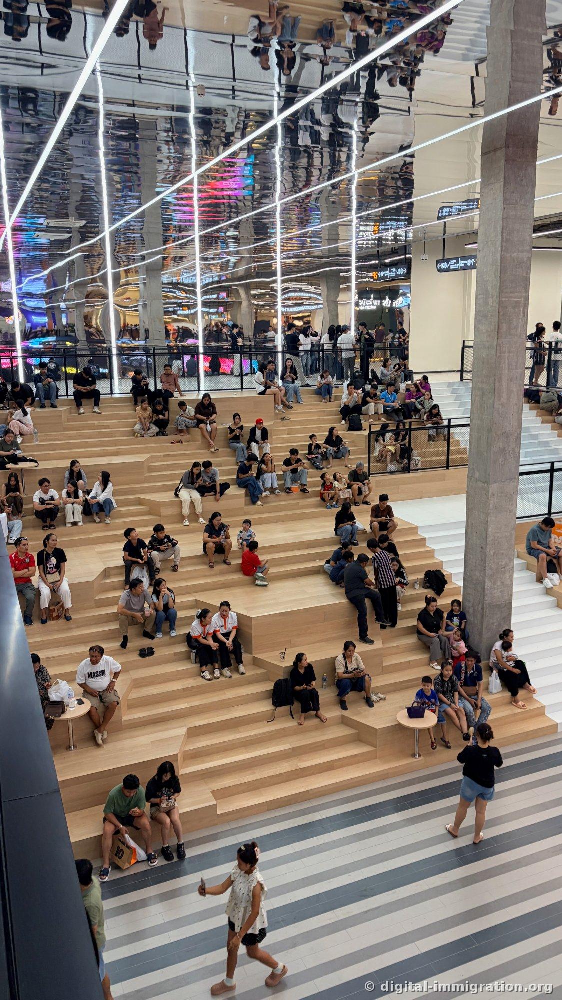 People resting on wooden stepped seating, ceiling is mirror-finished reflecting the scene below