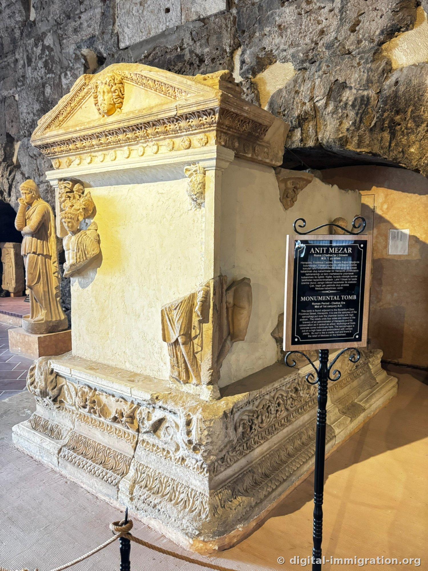 Large commemorative tomb with gabled roof lid carved with a Medusa face, black explanatory placard reading 'ANIT MEZAR / MONUMENTAL TOMB' placed beside it