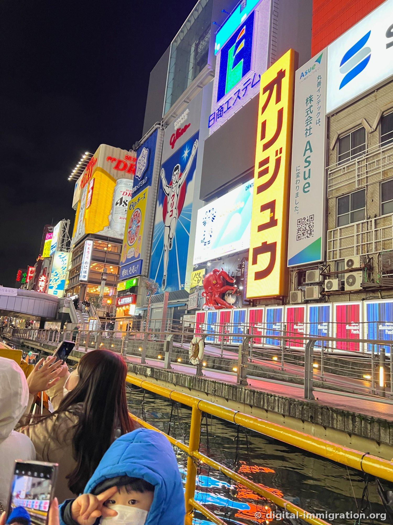 Enseigne Glico la nuit vue d'en bas depuis le bateau, enseigne Origen également visibles, avec des passagers prenant des photos avec leurs téléphones