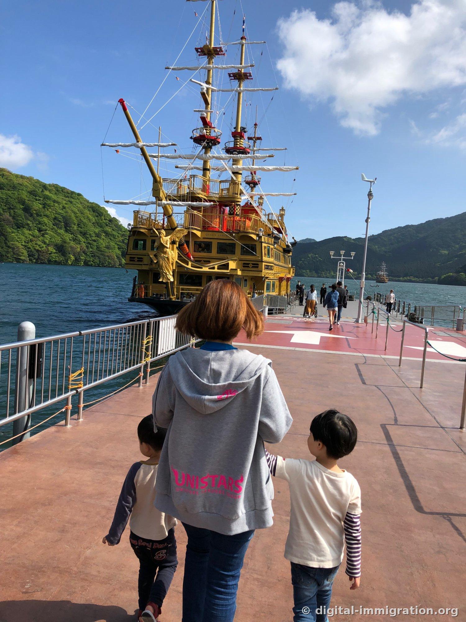 Deux enfants et un adulte marchant vers le bateau pirate doré sur le vaste ponton rouge, avec trois mâts et une statue dorée de déesse visible à l'avant du navire amarré