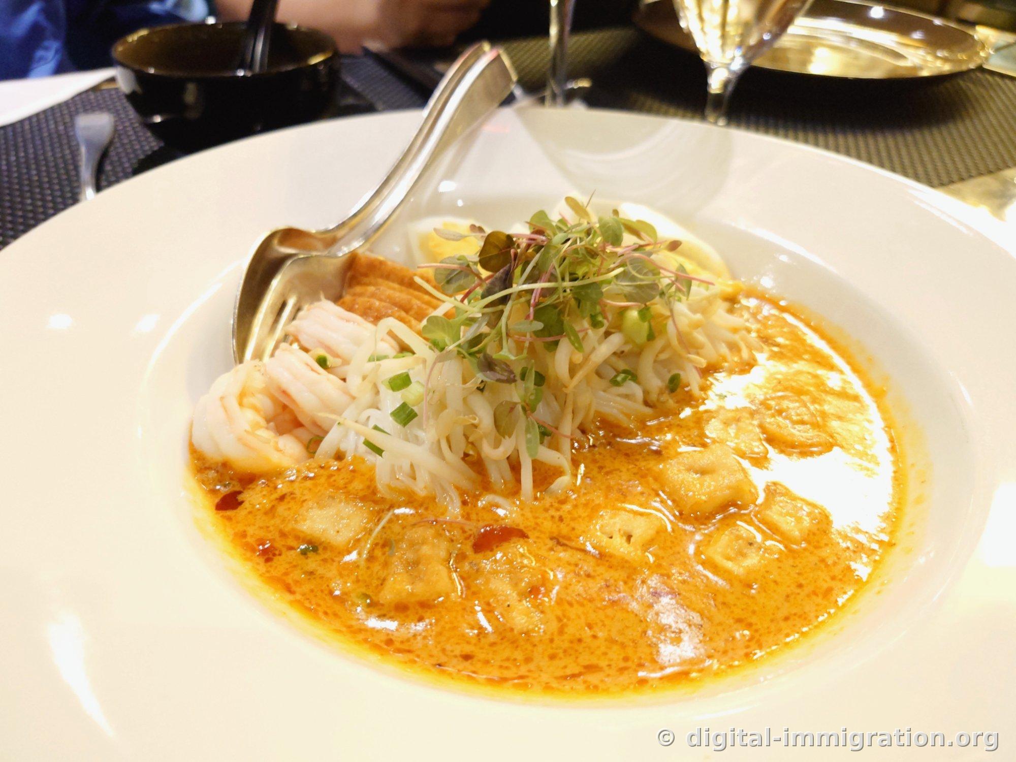 Coconut curry noodle soup on a white plate with orange broth, rice noodles, bean sprouts, shrimp, boiled egg, and fried tofu
