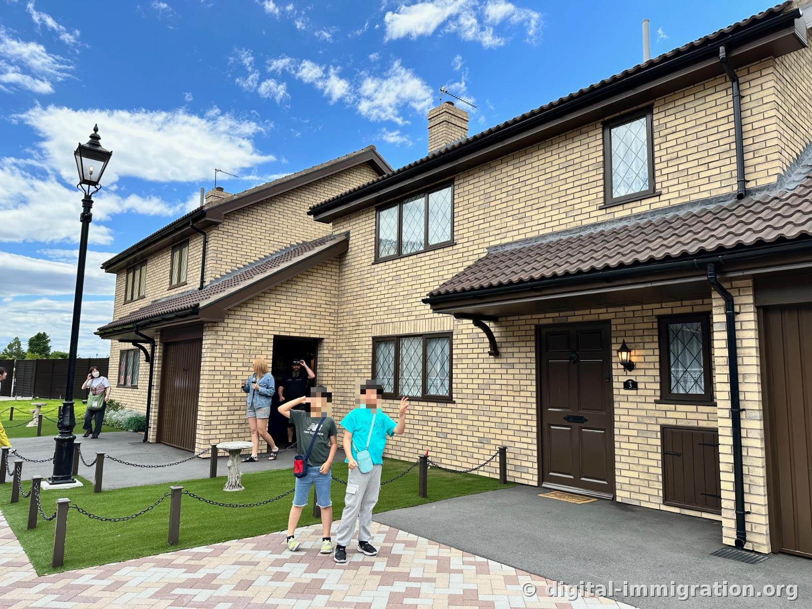 Two children posing in front of a yellow-brick house set under blue sky, with a black streetlamp and house number 3 on the adjacent home