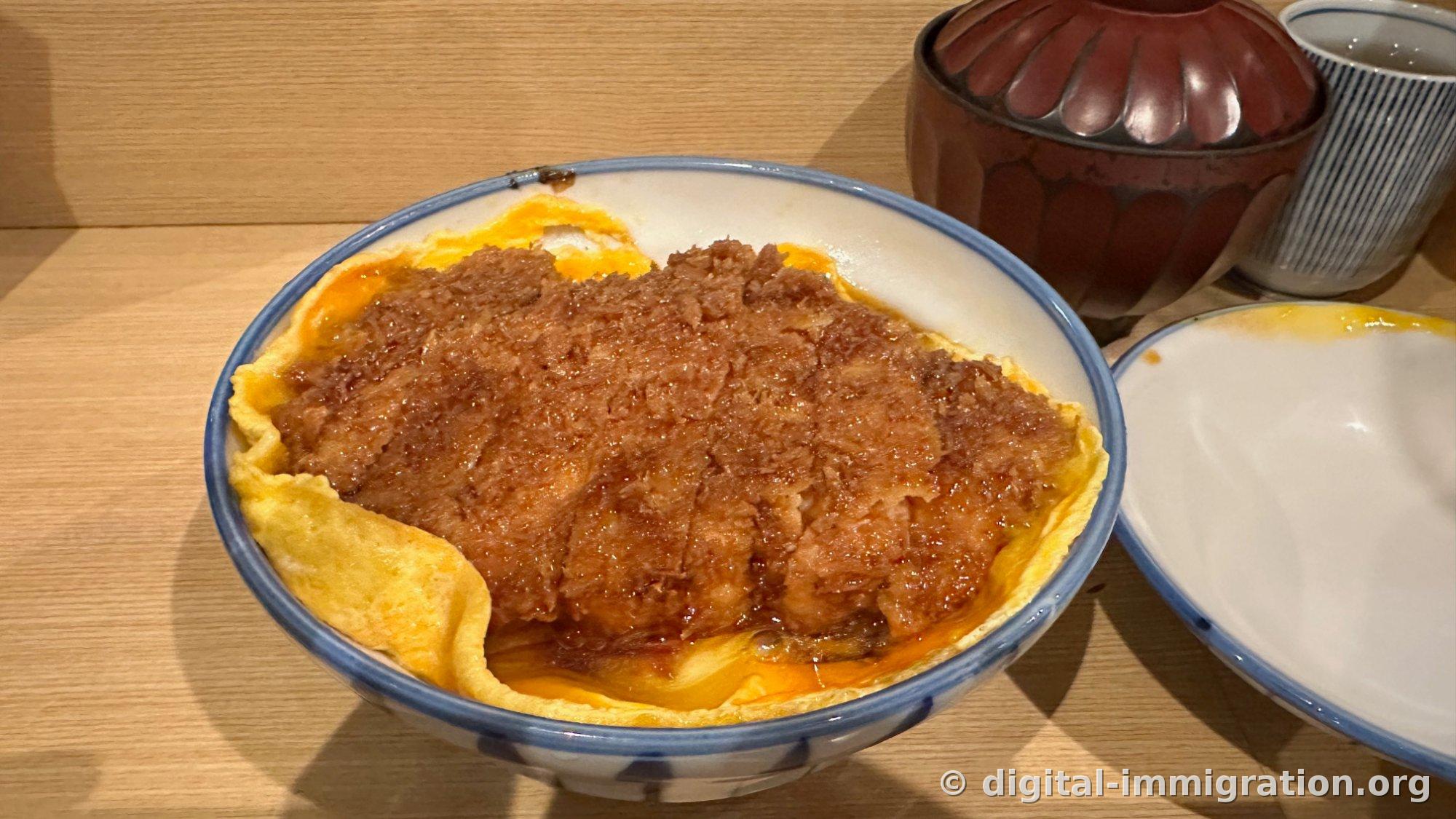 Une katsudon dans un bol à bordure bleue. L'omelette jaune enveloppe complètement le riz, et au-dessus repose la katsudon nappée de sauce. À côté se trouvent un bol en laque rouge et une tasse à rayures