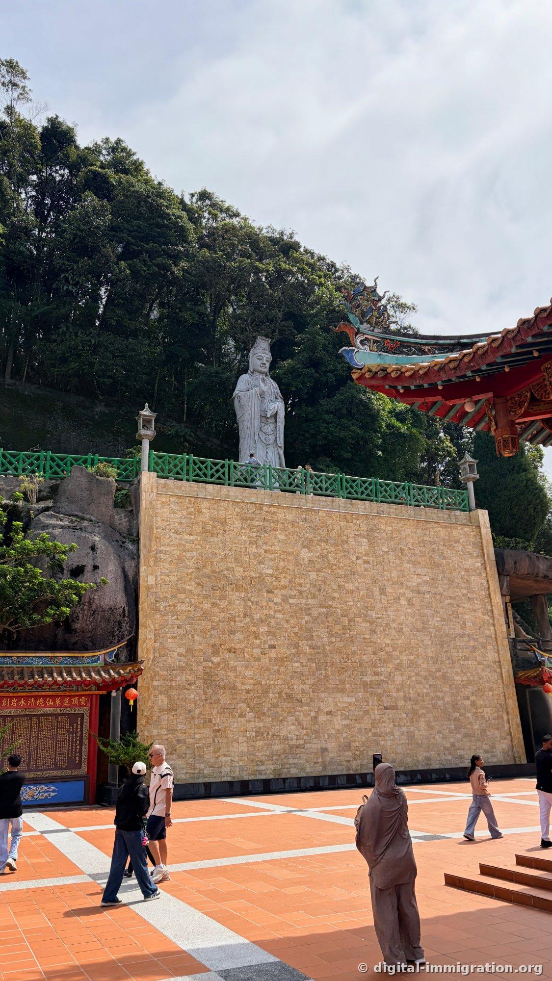 Une statue debout blanche de Guanyin sur un mur de pierre jaune, l'avant-toit d'un pavillon rouge à droite, des touristes marchant sur la place en carrelage orange