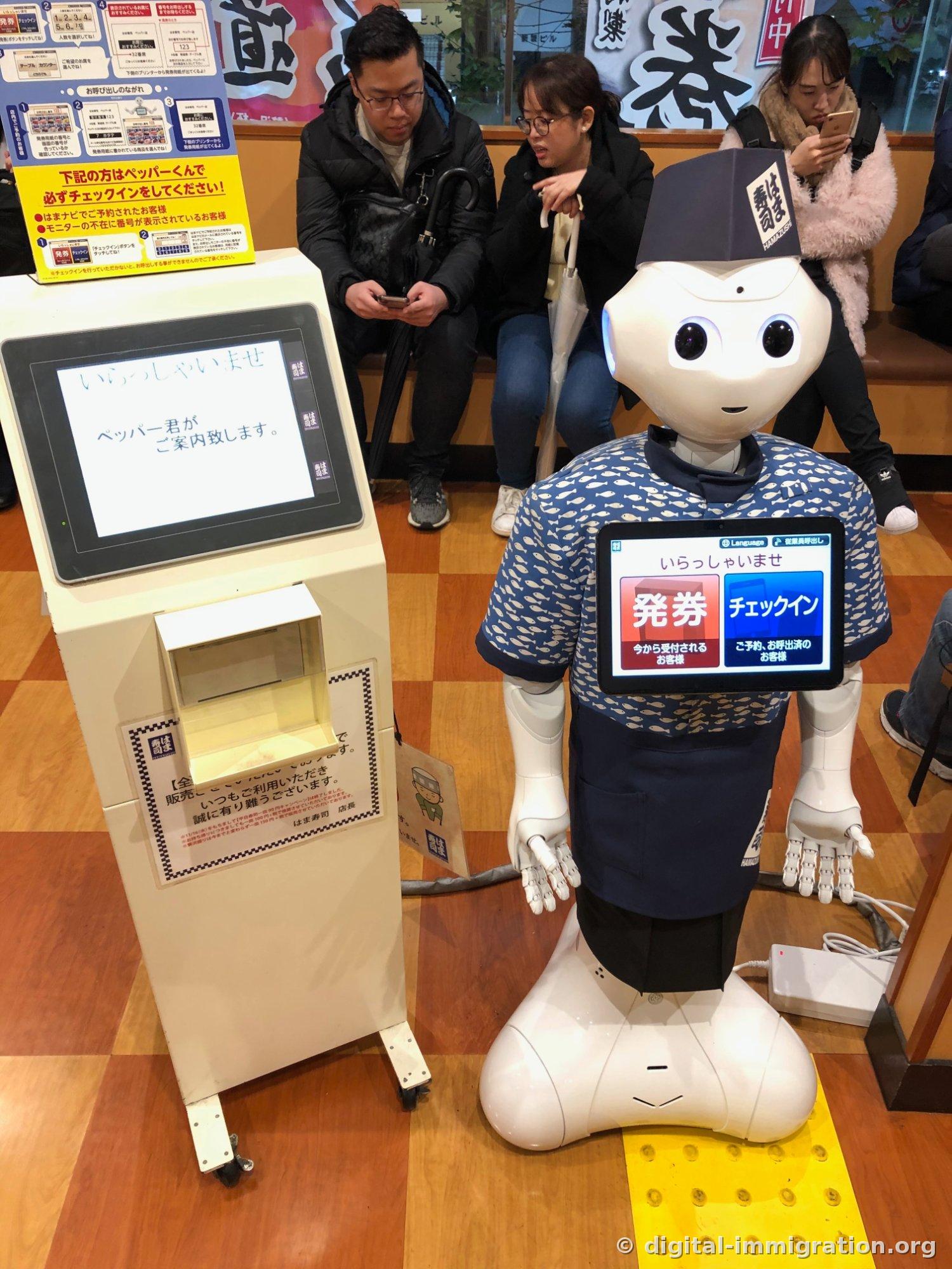 Un robot Pepper vêtu d'un tablier Hamasushi et d'un chapeau au logo Hamasushi affiche des boutons de délivrance de tickets et de check-in sur l'écran tactile de sa poitrine, et à côté se trouve une machine de délivrance de tickets avec un écran montrant いらっしゃいませ