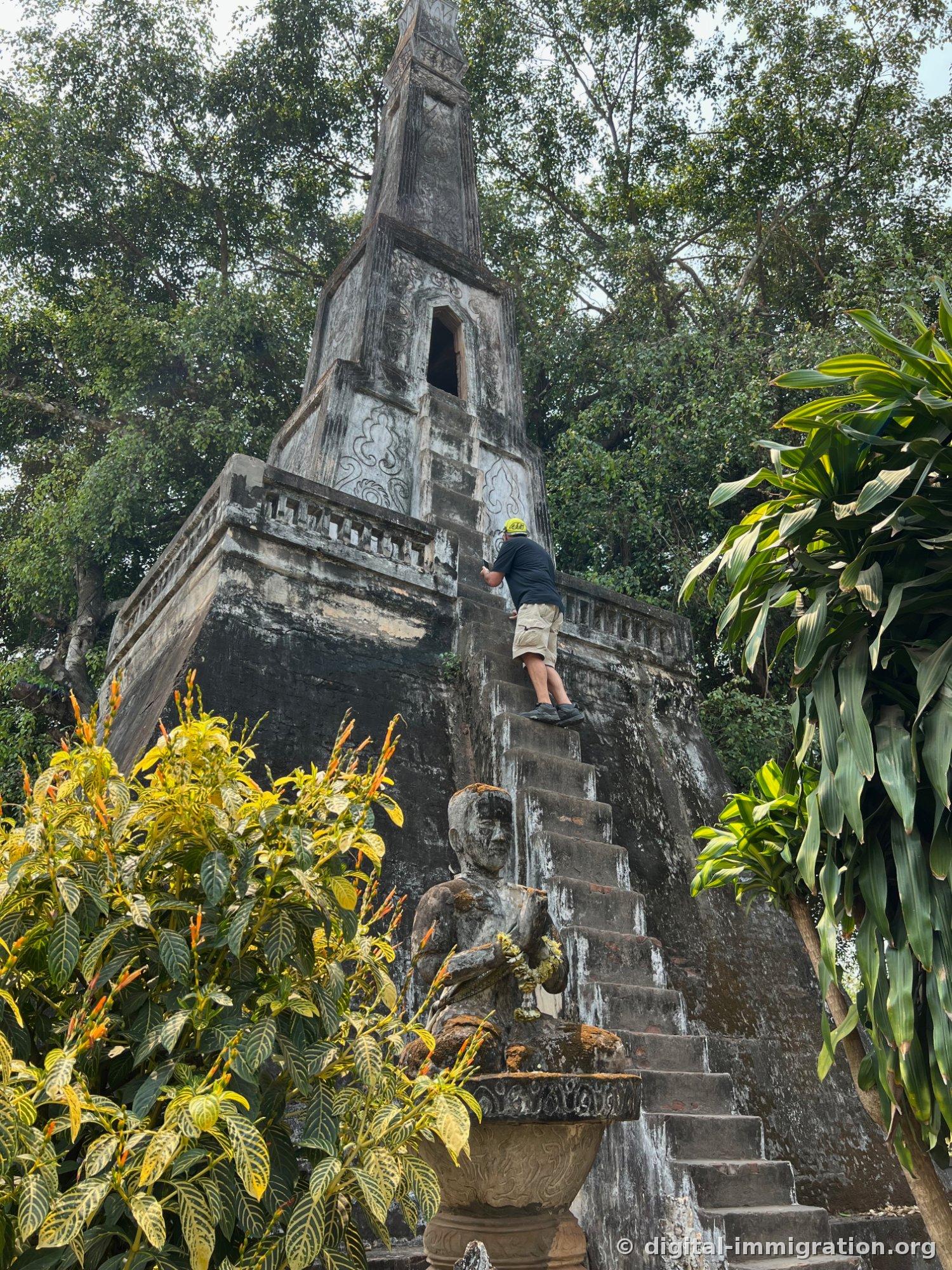Un homme au chapeau jaune montant un escalier étroit sans rampe sur une structure grise de type tour avec une statue assise en bas