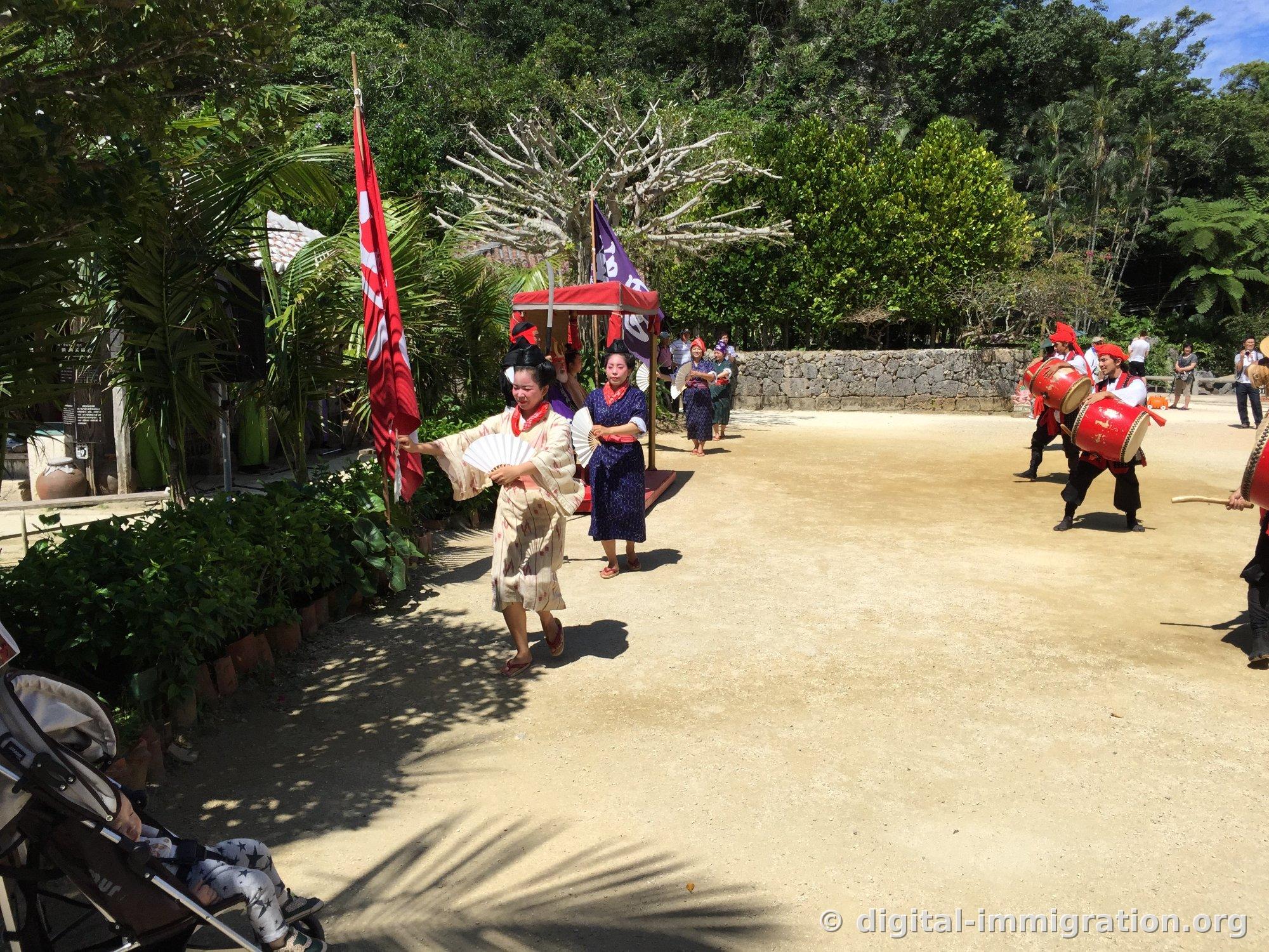 Une danseuse tenant un éventail blanc ouvert avance, tandis qu'à l'arrière, le palanquin rouge et les drapeaux sont visibles