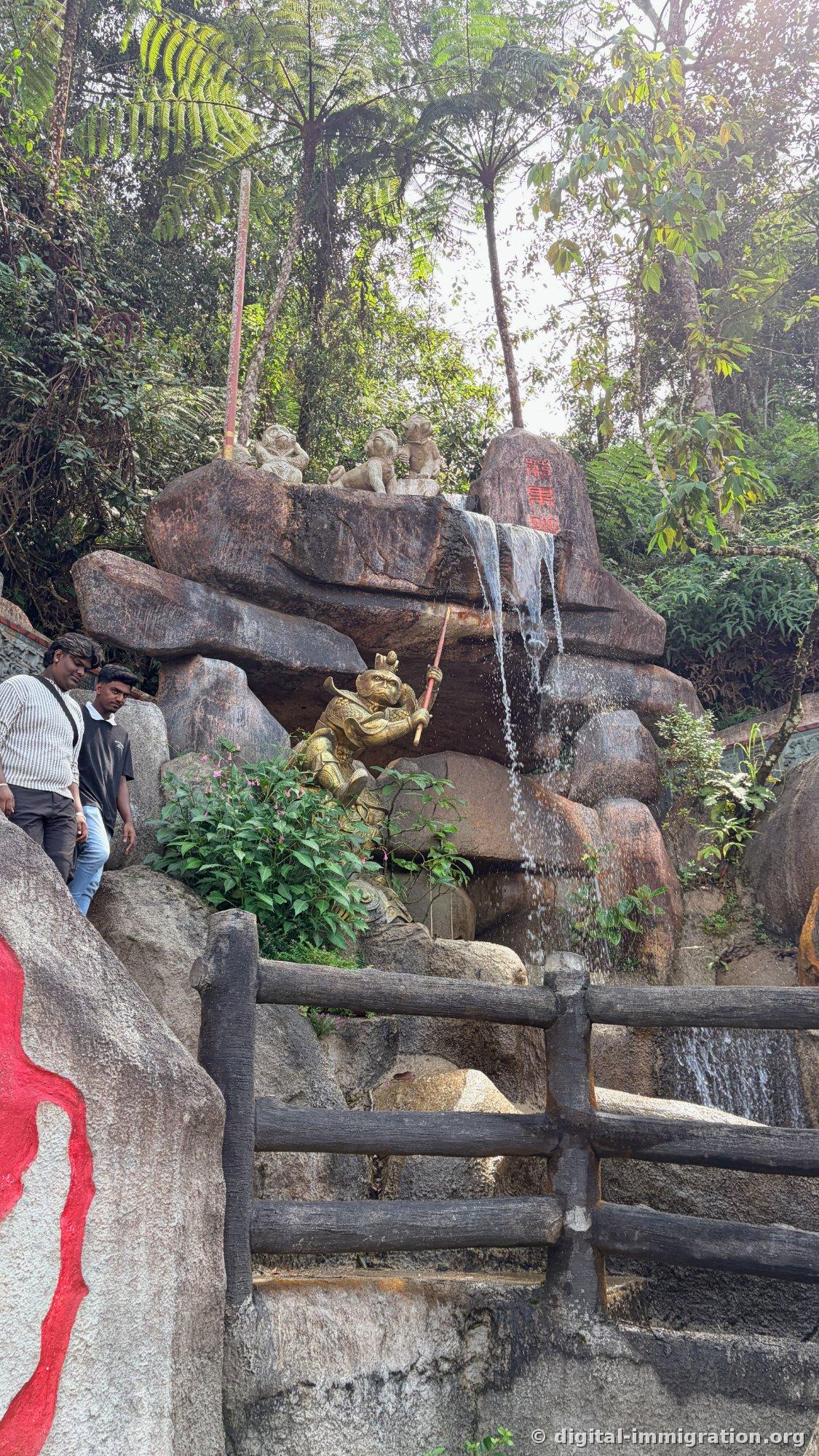 Cascade artificielle entre les rochers avec une sculpture dorée tenant une lance et des figurines de singes sur les rochers, deux hommes descendant les escaliers en rondins derrière