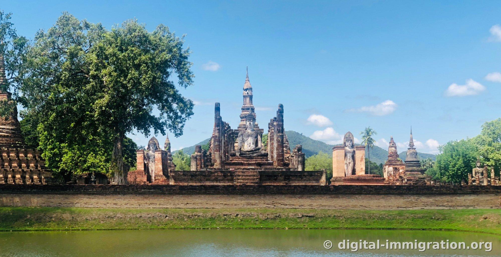 Wat Mahathat seen from across the moat, with the lotus-bud spire in the center, standing Buddha on the left, reclining Buddhas in niches on both sides, and a mountain ridge in the back
