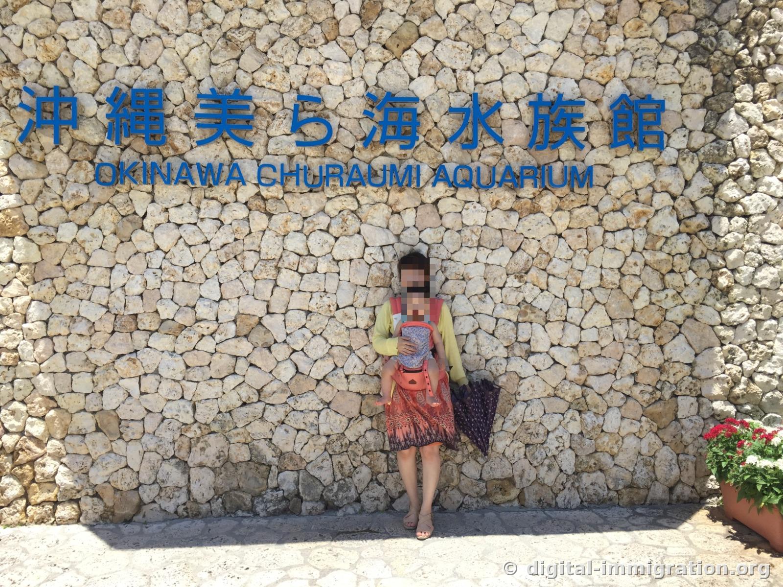 琉球石灰岩墙上用蓝色字体写着冲绳美丽海水族馆 OKINAWA CHURAUMI AQUARIUM，一个背着婴儿背带的人站在前面