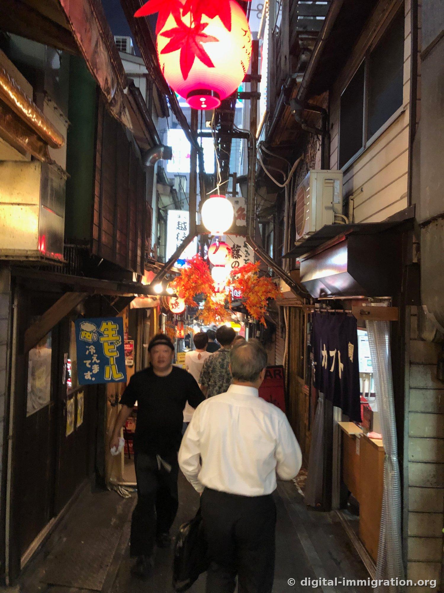 Une ruelle étroite avec des lanternes rouges pendent bas, un panneau de bière fraîche en bleu et des passants