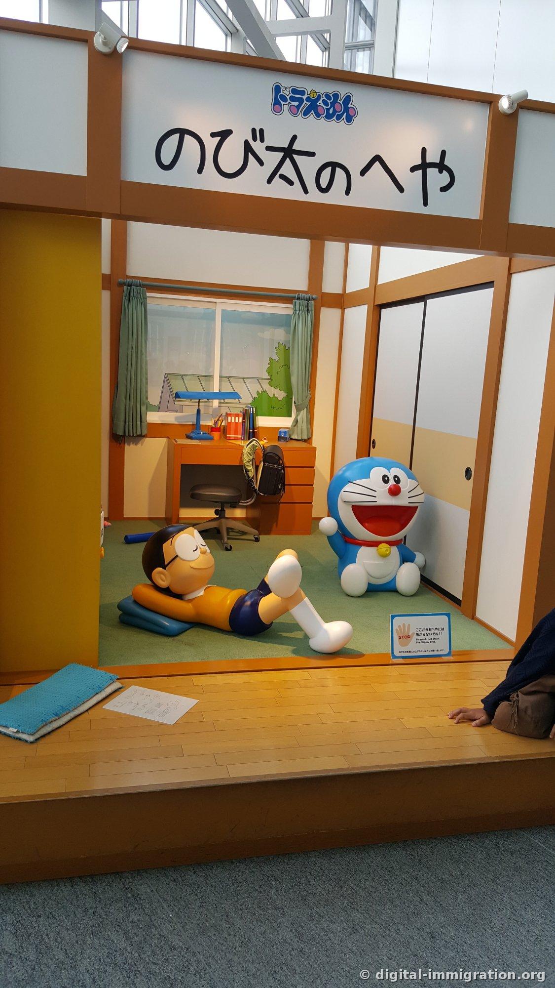 Enseigne 'La chambre de Nobita' au-dessus d'une pièce en tatami reconstituée à taille réelle avec une figurine de Suneo dormant sur le sol et Doraemon assis à côté, avec un panneau d'interdiction d'accès à l'avant