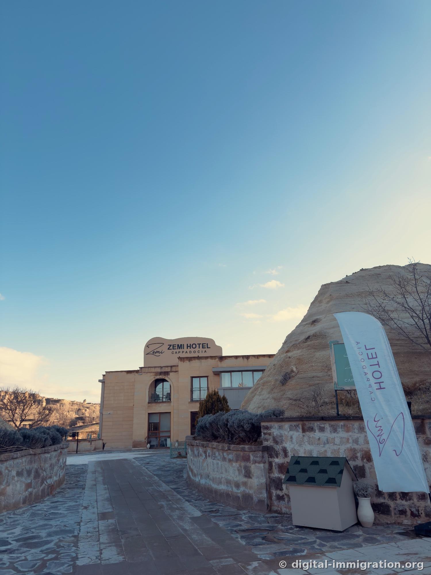 Sous un ciel dégagé, un bâtiment en pierre beige avec un panneau 'ZEMI HOTEL CAPPADOCIA', un énorme rocher de tuf attaché à droite, un mur en pierre et une bannière dans la cour de devant