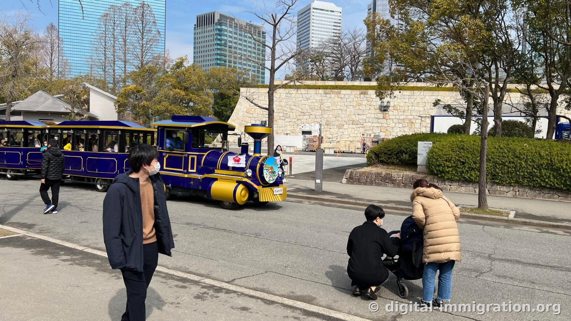 Le petit train touristique Sumire-go peint en violet-noir et doré traversant une route du parc du château d'Osaka avec ses wagons qui le suivent, et une famille qui l'observe depuis le bord