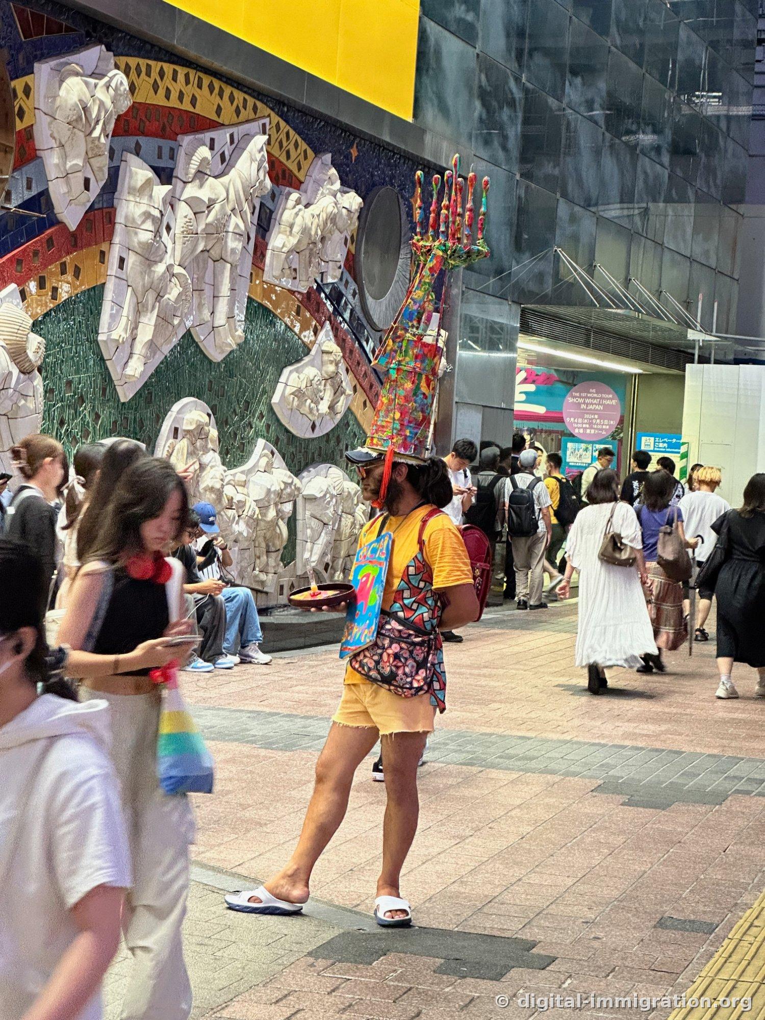 頭にカラフルな塔の装飾を乗せ、黄色いTシャツにスリッパ姿で歩く人、後ろに白い浮き彫りの壁画が見える