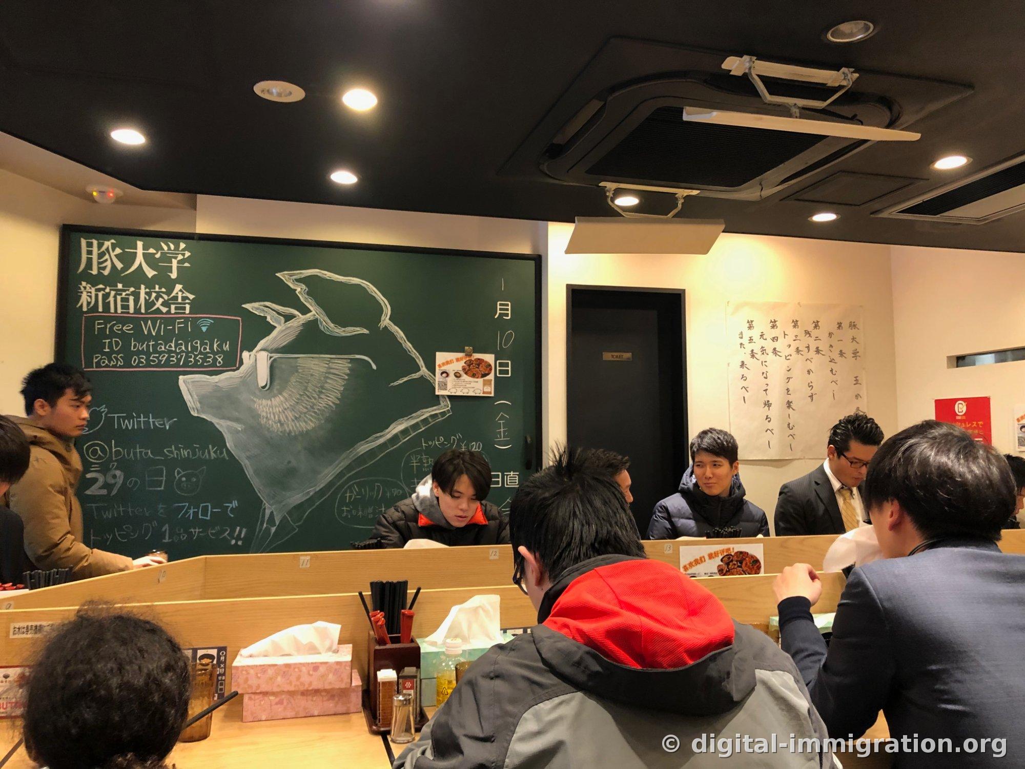 L'intérieur du restaurant avec son tableau noir vert affichant « 豚大学 新宿校舎 » et un dessin d'un cochon portant des lunettes, l'ID Wi-Fi gratuit et un compte Twitter écrit à la craie, et le comptoir rempli de clients