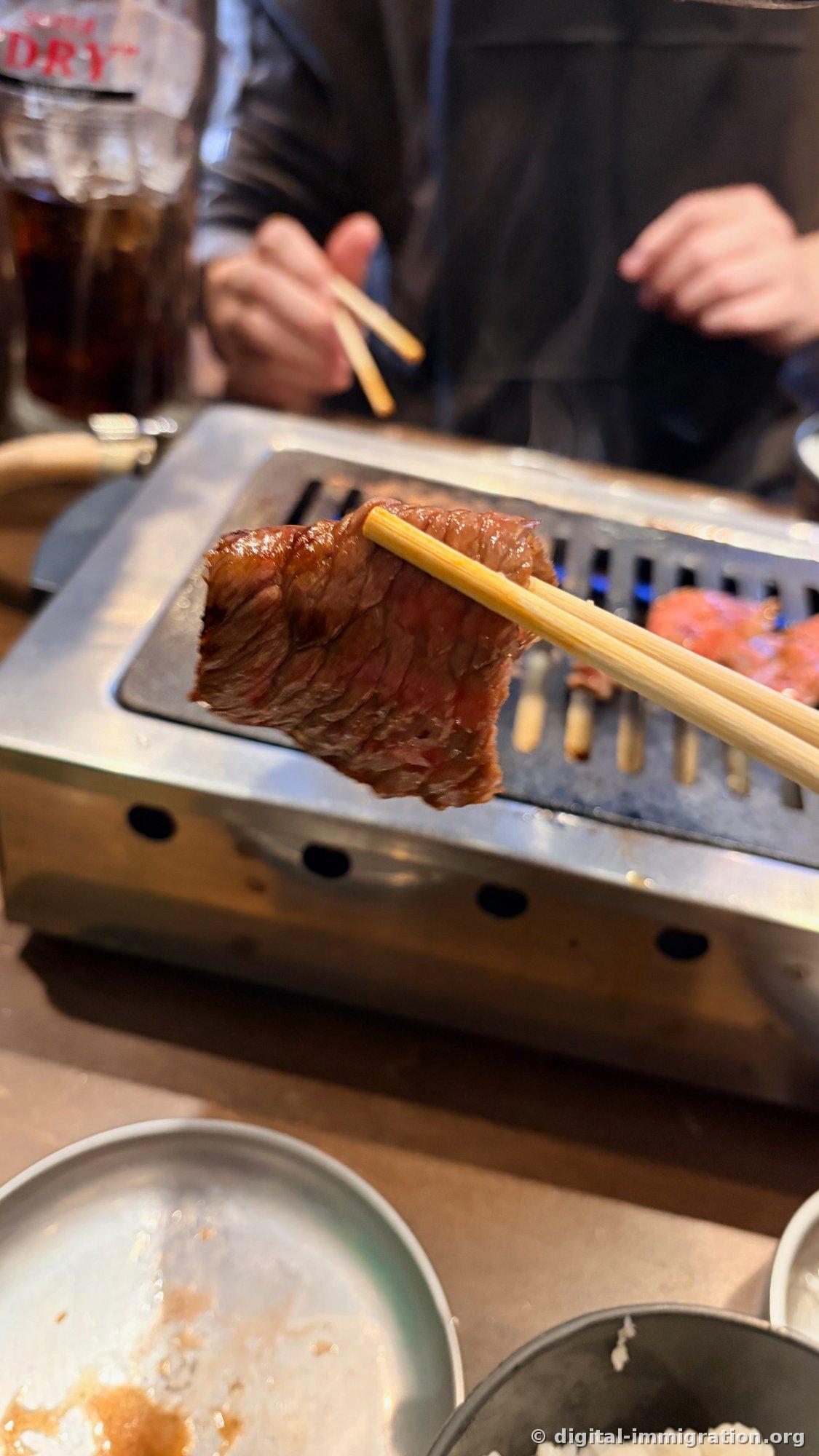 A slice of grilled beef with a brown exterior and pink interior lifted with chopsticks; oolong tea glass and an apron-wearing companion visible in the background