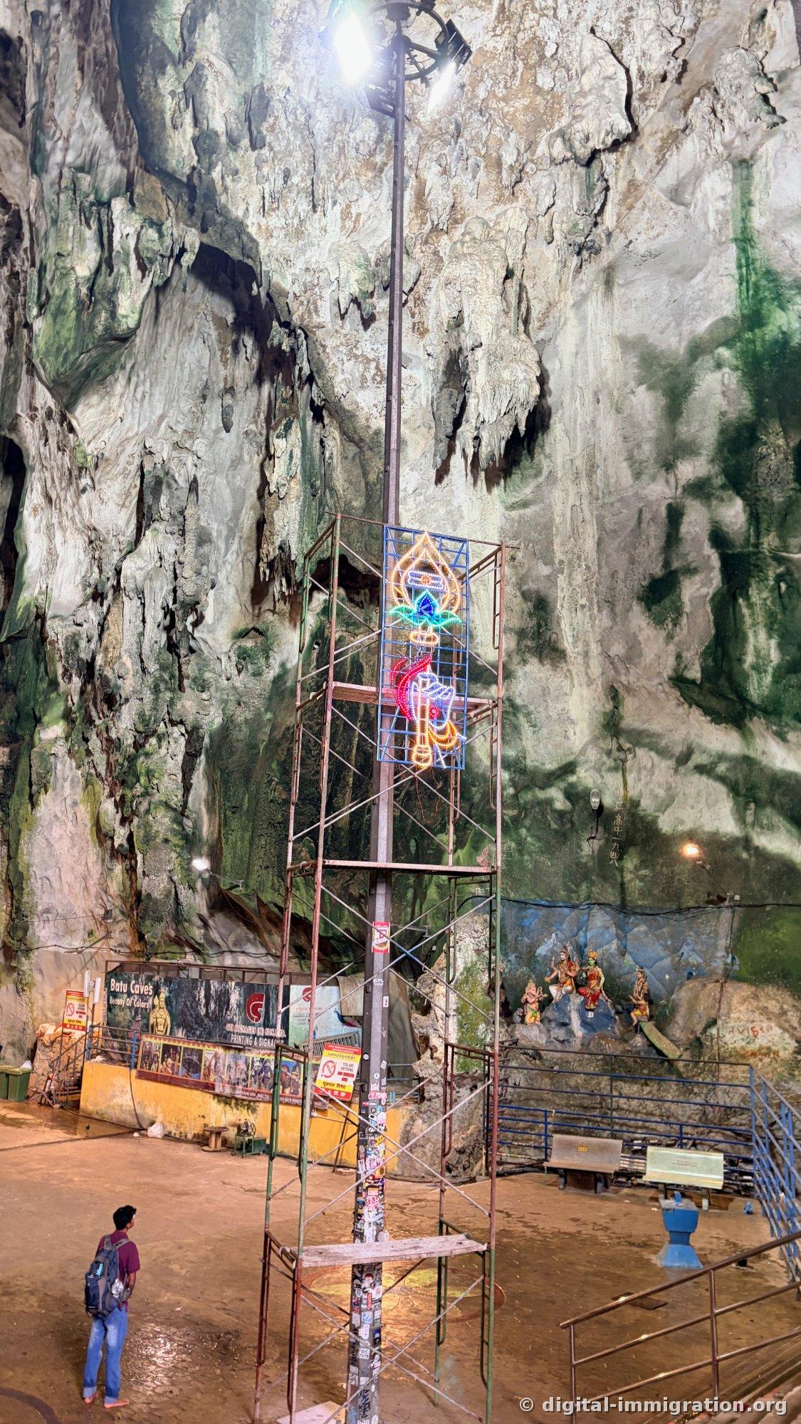 洞窟の中の鉄製の足場に取り付けられたネオン装飾と高い照明塔、壁面に'Batu Caves'の横幕とヒンドゥーの神像の彫刻があり、バックパックを背負った人が見上げている