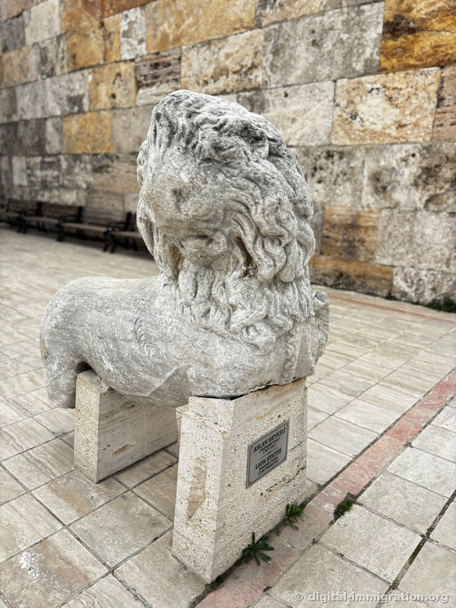 Intact marble lion statue resting on outdoor stone pedestal with stone wall and benches visible behind, nameplate reading 'ASLAN HEYKELI / LION STATUE' attached to base