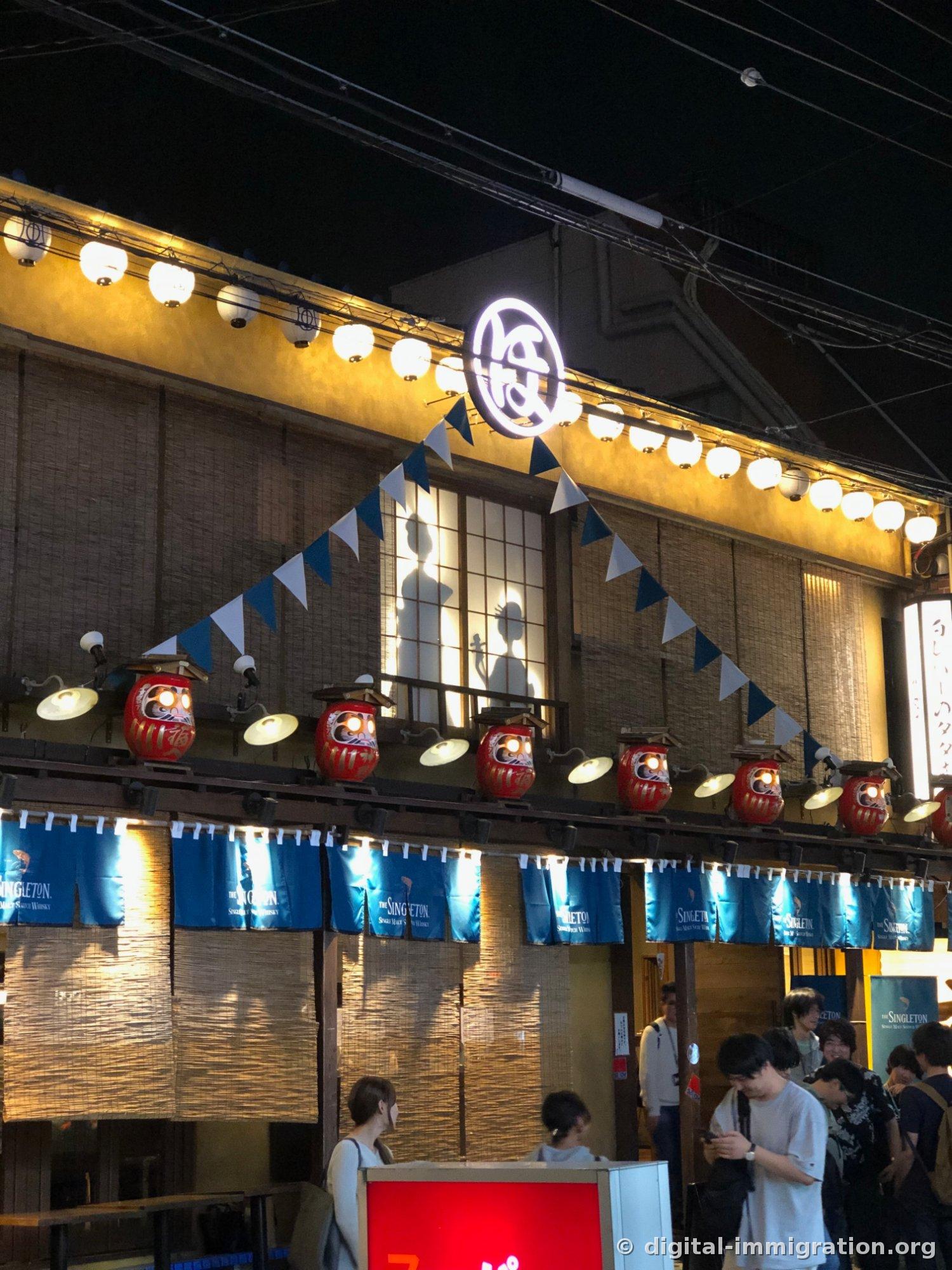 红色达摩灯笼和白色纸灯笼沿着屋檐挂起，店铺正面。蓝色暖帘垂下，和纸门上映出人影