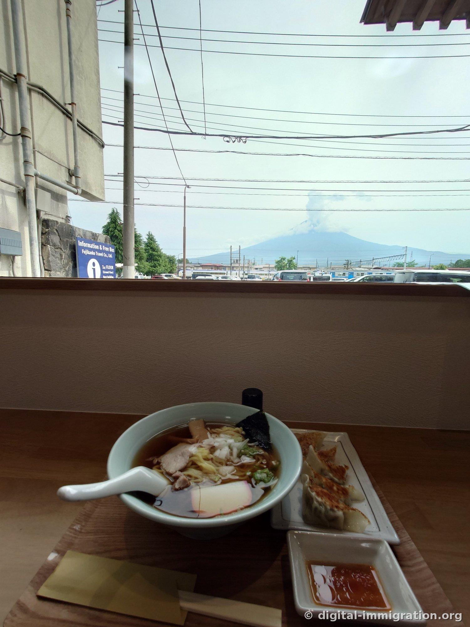 窓際のカウンター席に置かれたラーメンと餃子の向こうに、富士山の頂上から雲が白い煙のように立ち上っている様子