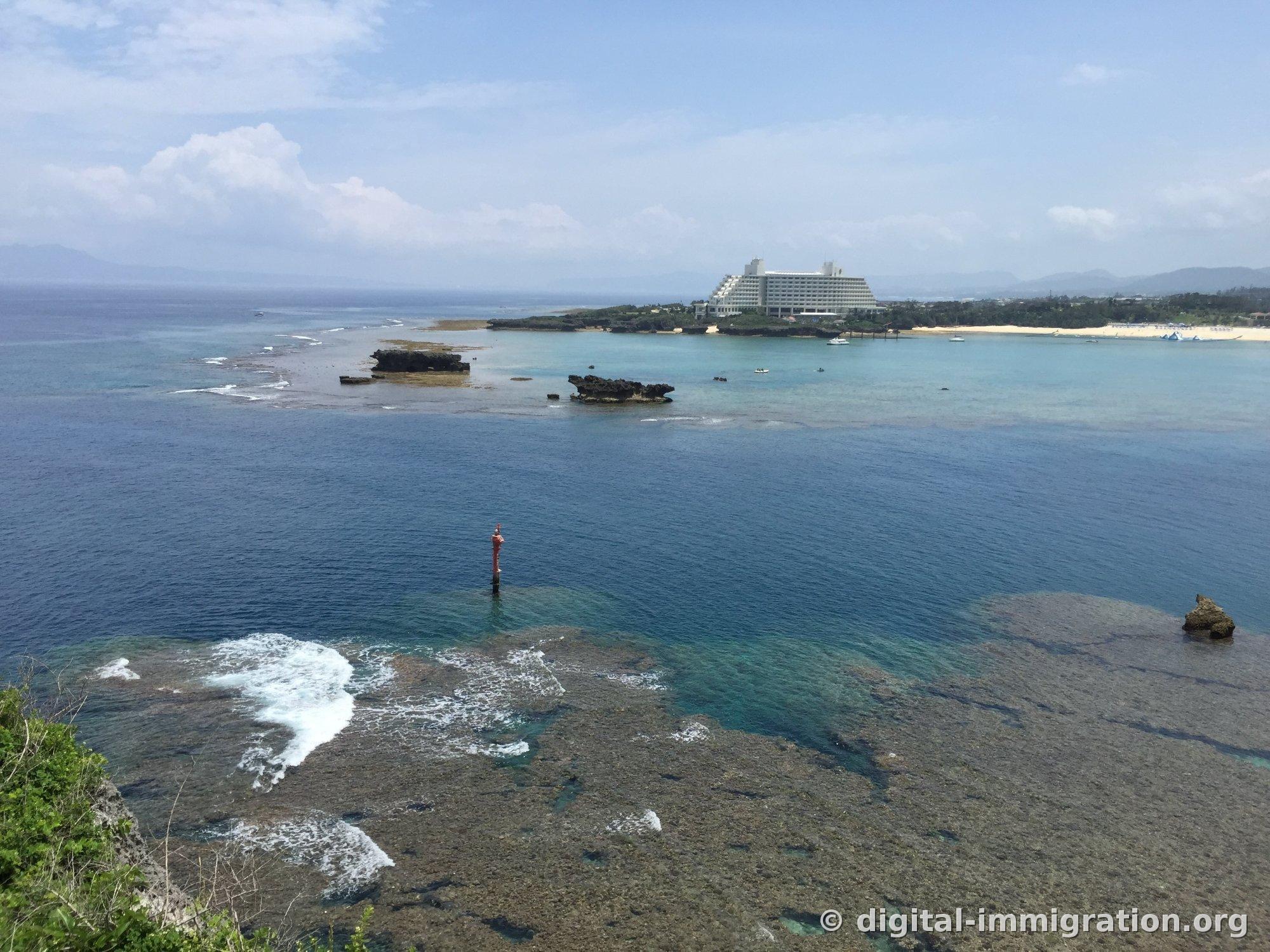 海の真ん中に赤い航路標識柱が立っており、その向こう側の岬の上に階段状の外観の白いリゾートホテルが見える万座毛の眺め