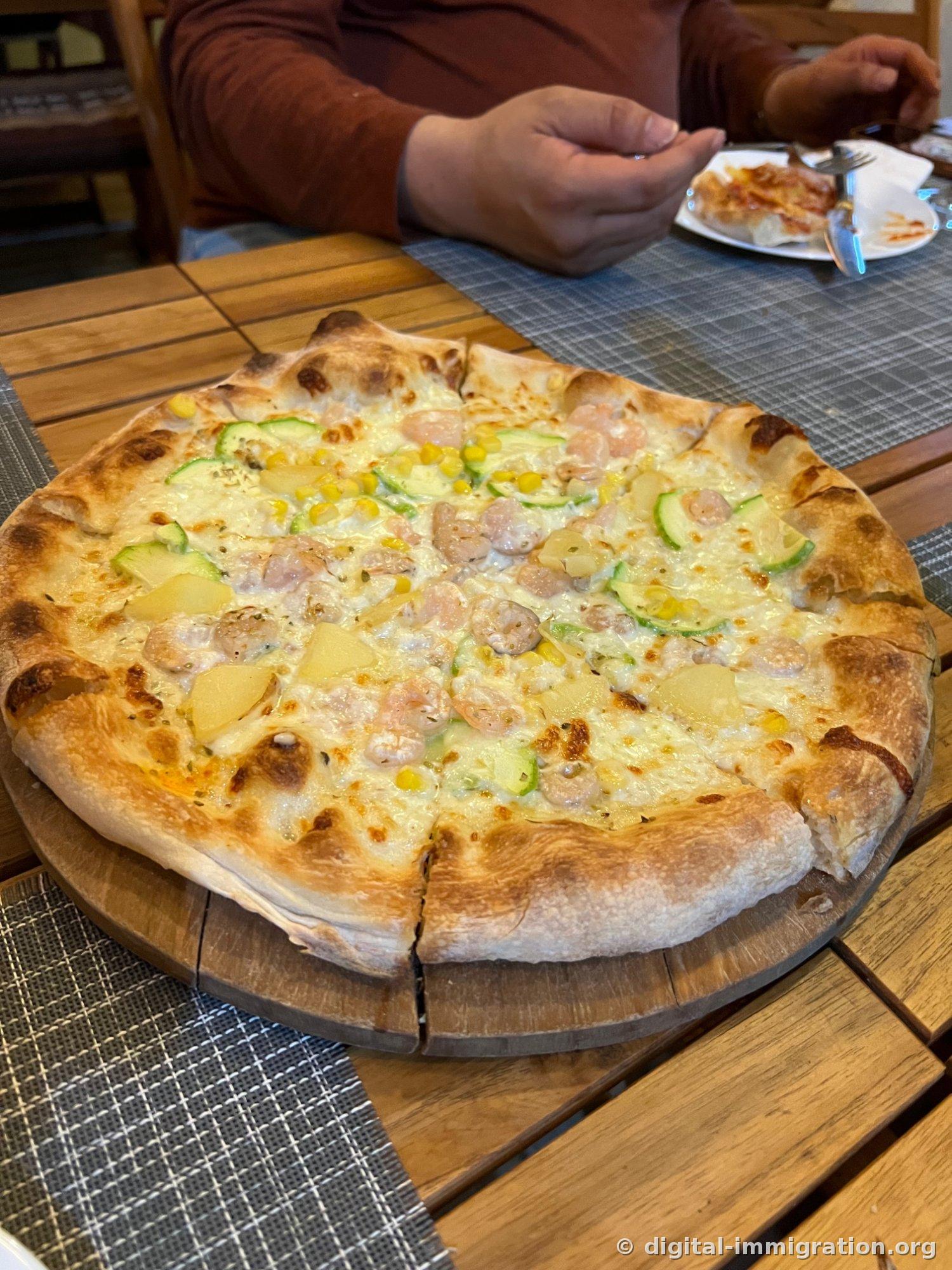 A white pizza on a wooden board. Topped with shrimp, potato, thinly sliced zucchini, and corn, with the crust edges puffed up and browned