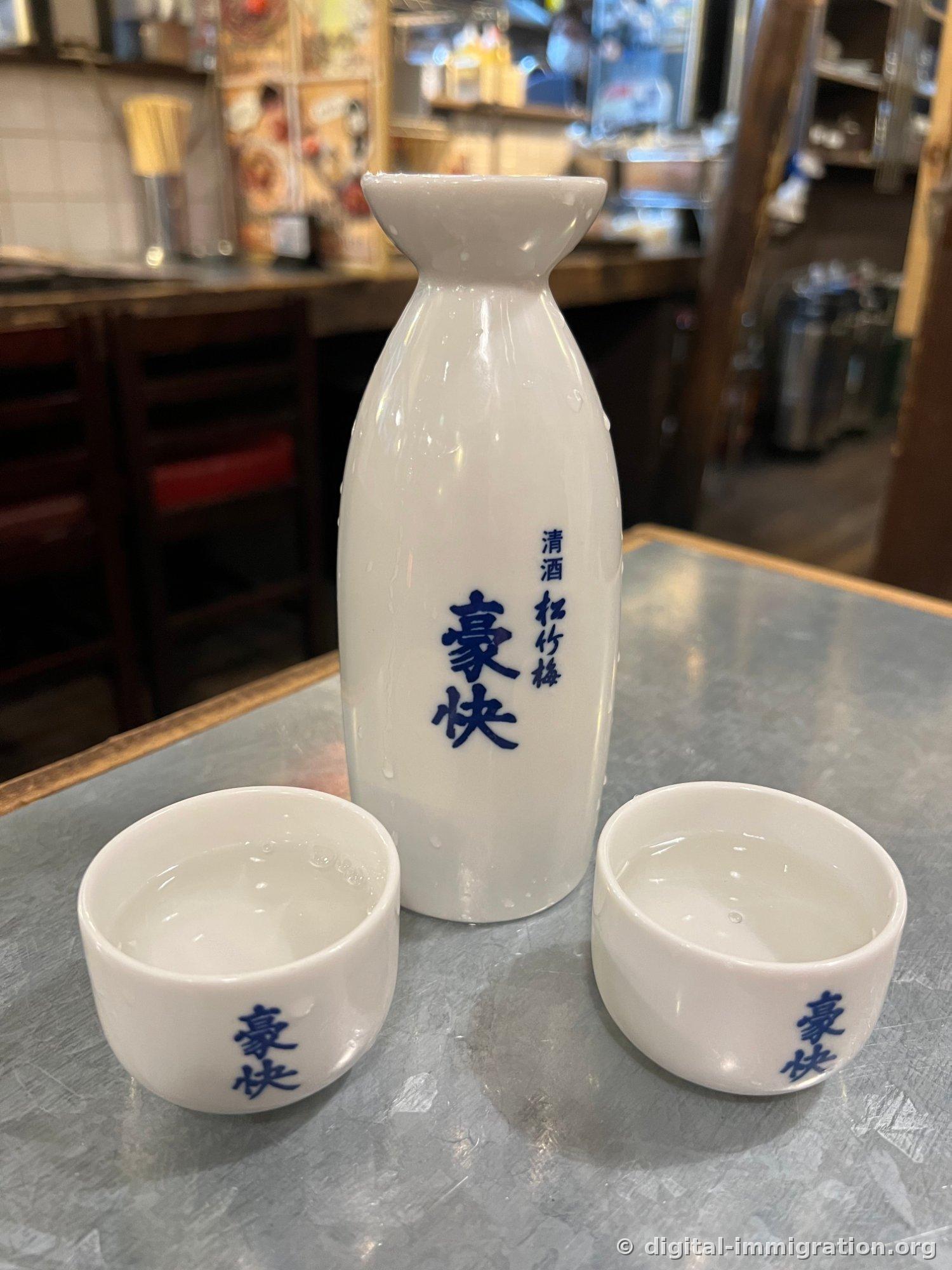 鉄製テーブルの上に置かれた白い陶製の酒瓶と杯二つ。青い字で「清酒 松竹梅 豪快」と書いてあり、瓶に水滴がついている