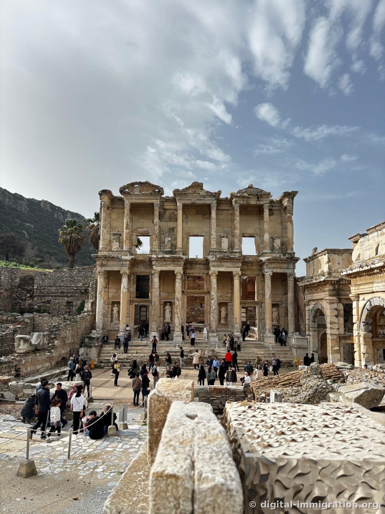 Full view of the Celsus Library two-story facade overlooked from a high vantage point above, with tourists gathered in the square below and the Mazeus Mithridates Gate on the right