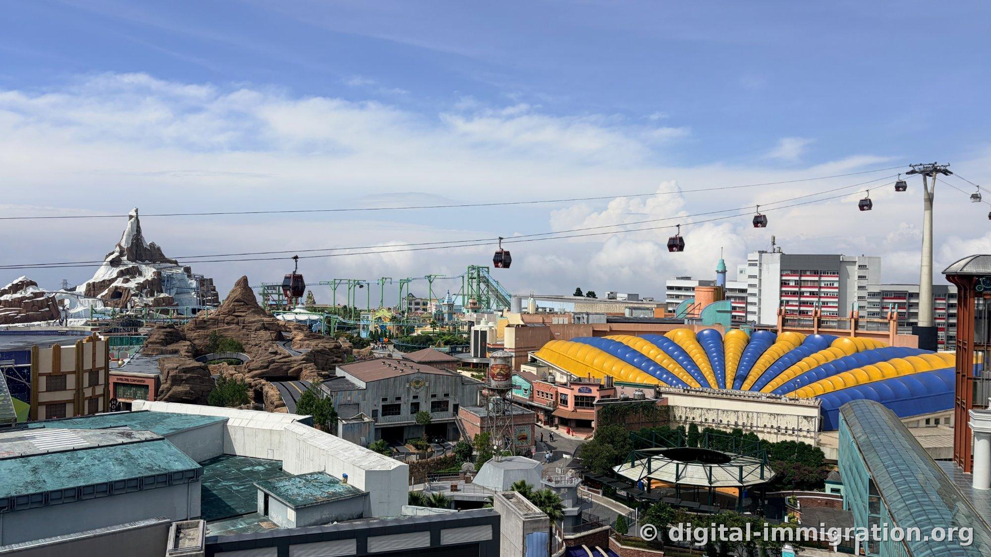 Red cable car cabins passing over the park with blue and yellow striped air dome and large residential building behind