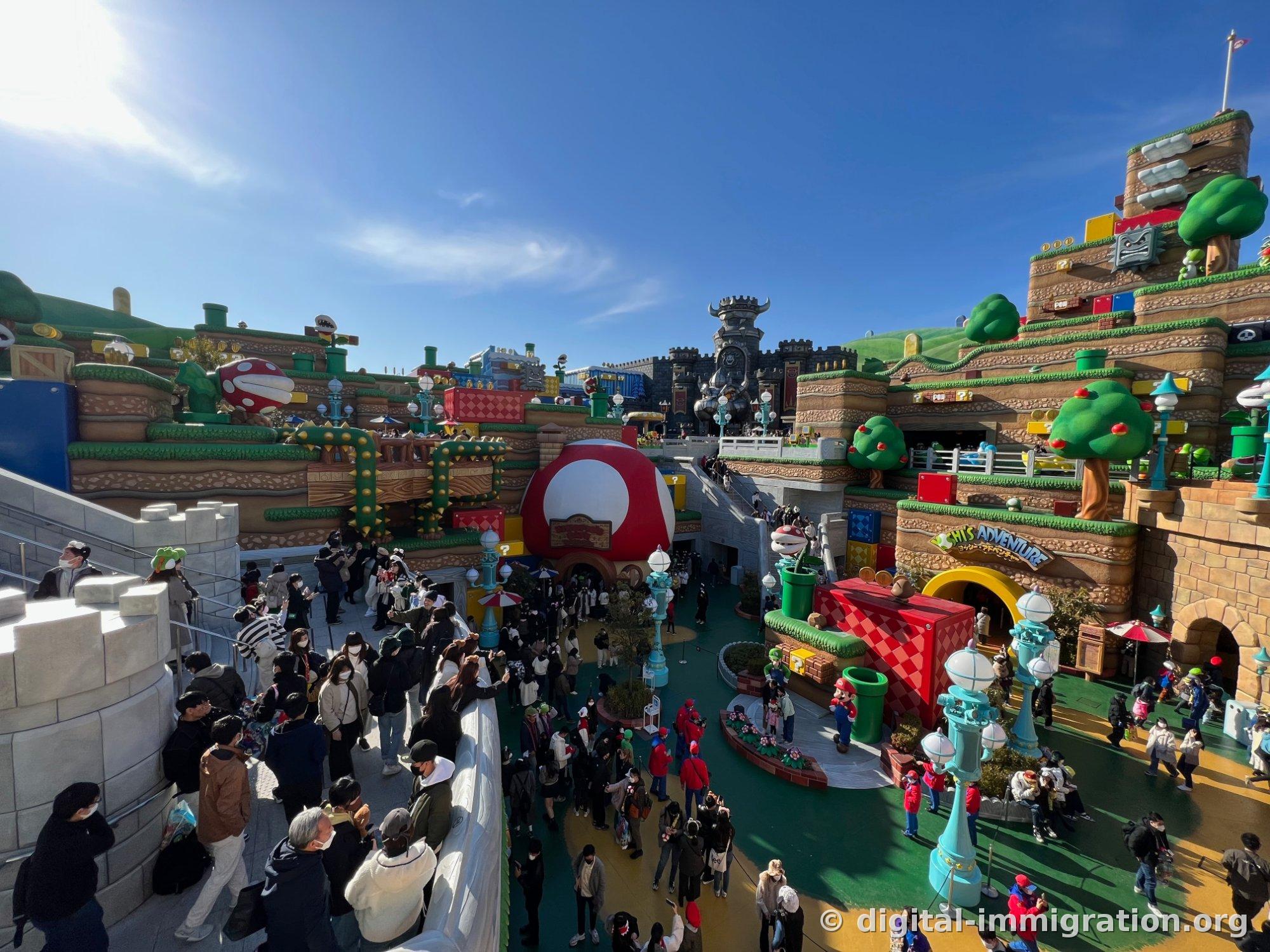 Super Nintendo World plaza viewed from above. The red mushroom-shaped Toad House and Bowser's Castle, with masked visitors filling the green floor
