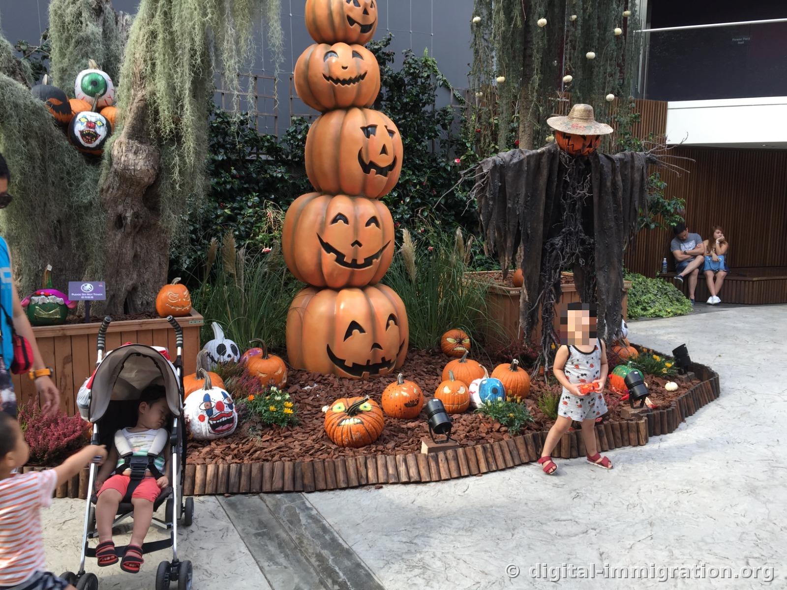 A tower of five stacked jack-o'-lanterns and a scarecrow in rags, with various painted pumpkins scattered on the ground and a child sleeping in a stroller